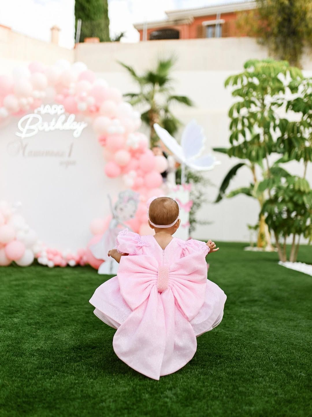 Back view of a baby girl walking toward a massive pink and white balloon arch setup at an outdoor event venue.