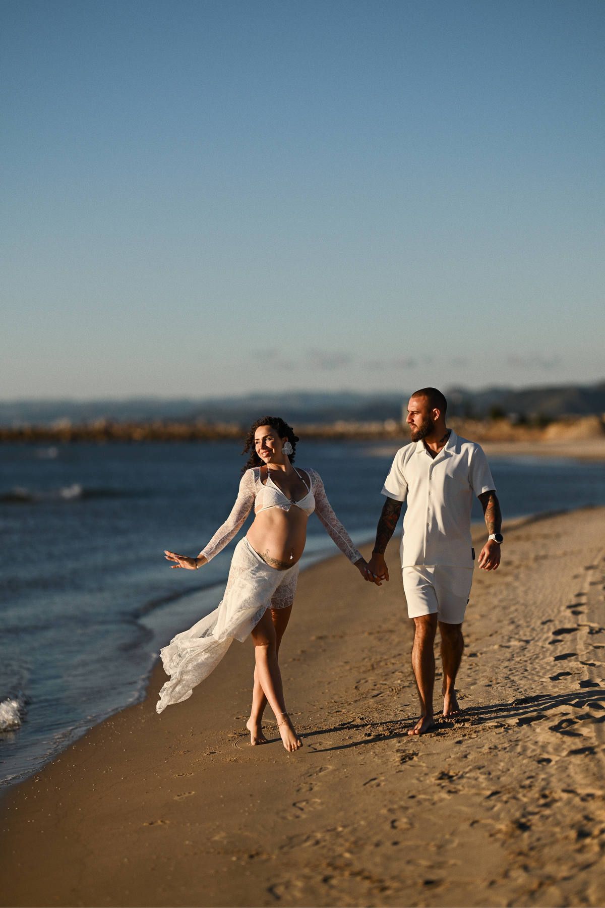 A joyful pregnant woman walking hand-in-hand with her partner along the shoreline during a sunlit beach maternity session.