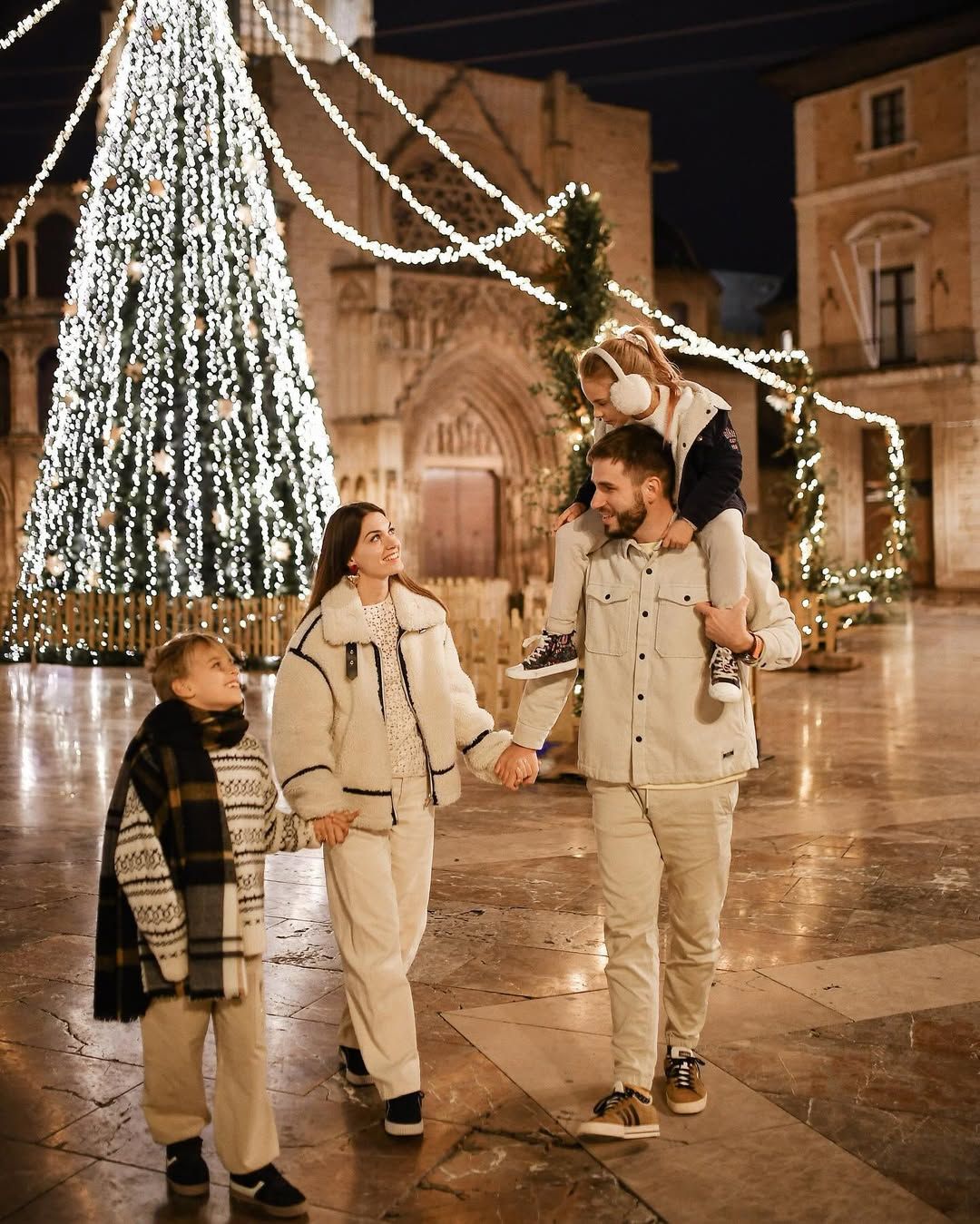 Family enjoying a festive evening walk in Valencia, with the father carrying his daughter on his shoulders by a Christmas tree