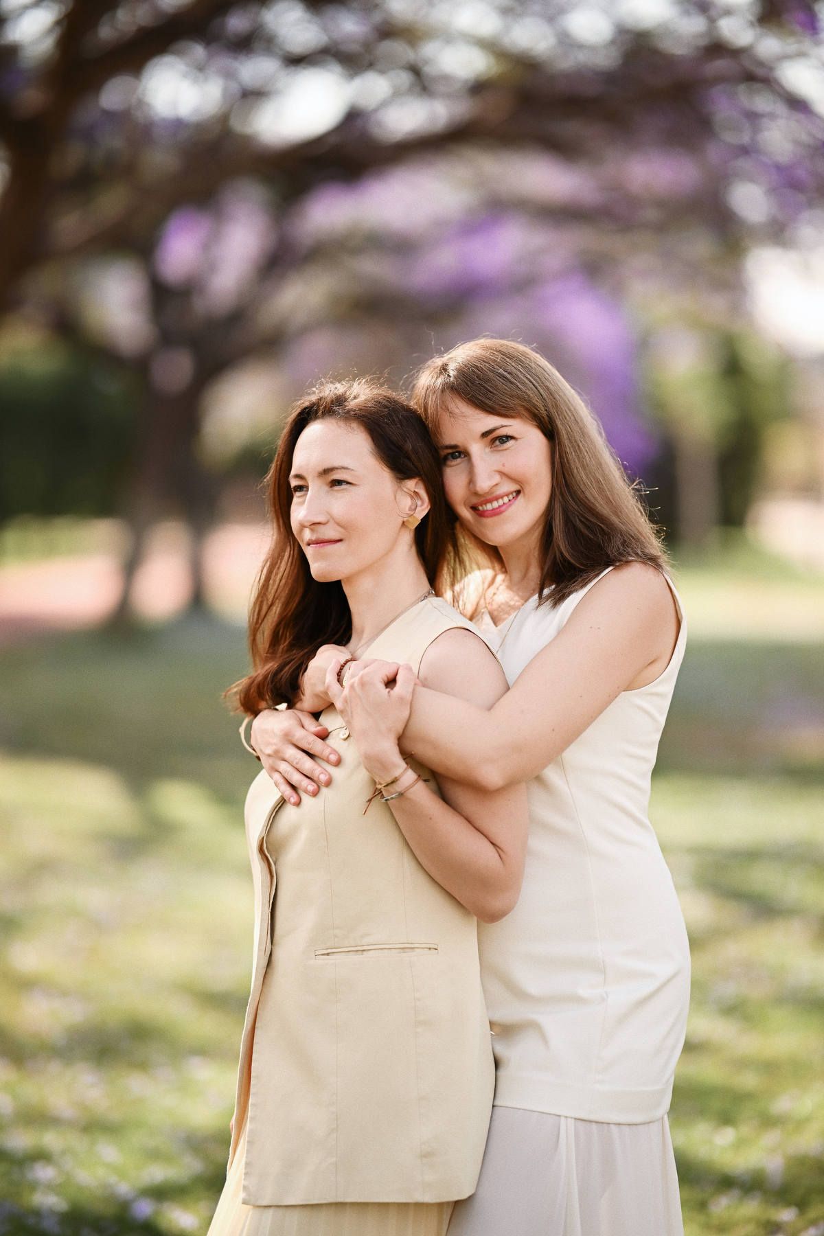 One sister lovingly embracing the other from behind, posing elegantly on a green lawn with purple trees in the background.