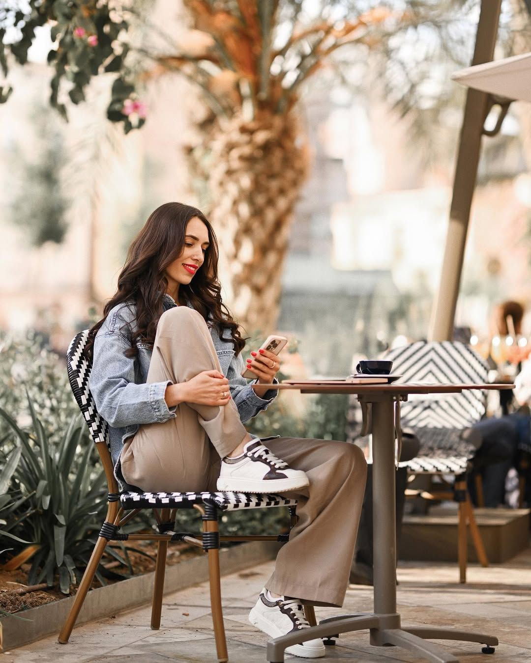 Smiling woman checking her phone at a cafe table with palm trees in Valencia. Casual lifestyle branding portrait