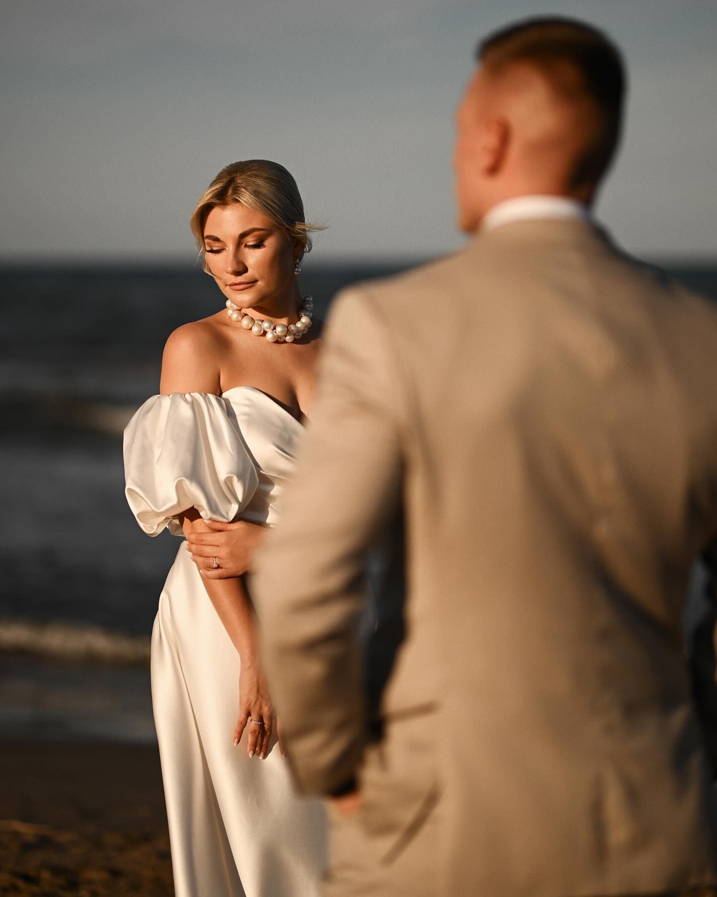 An editorial wedding portrait of a beautiful bride in a white dress standing by the sea, viewed past the groom's shoulder at golden hour.
