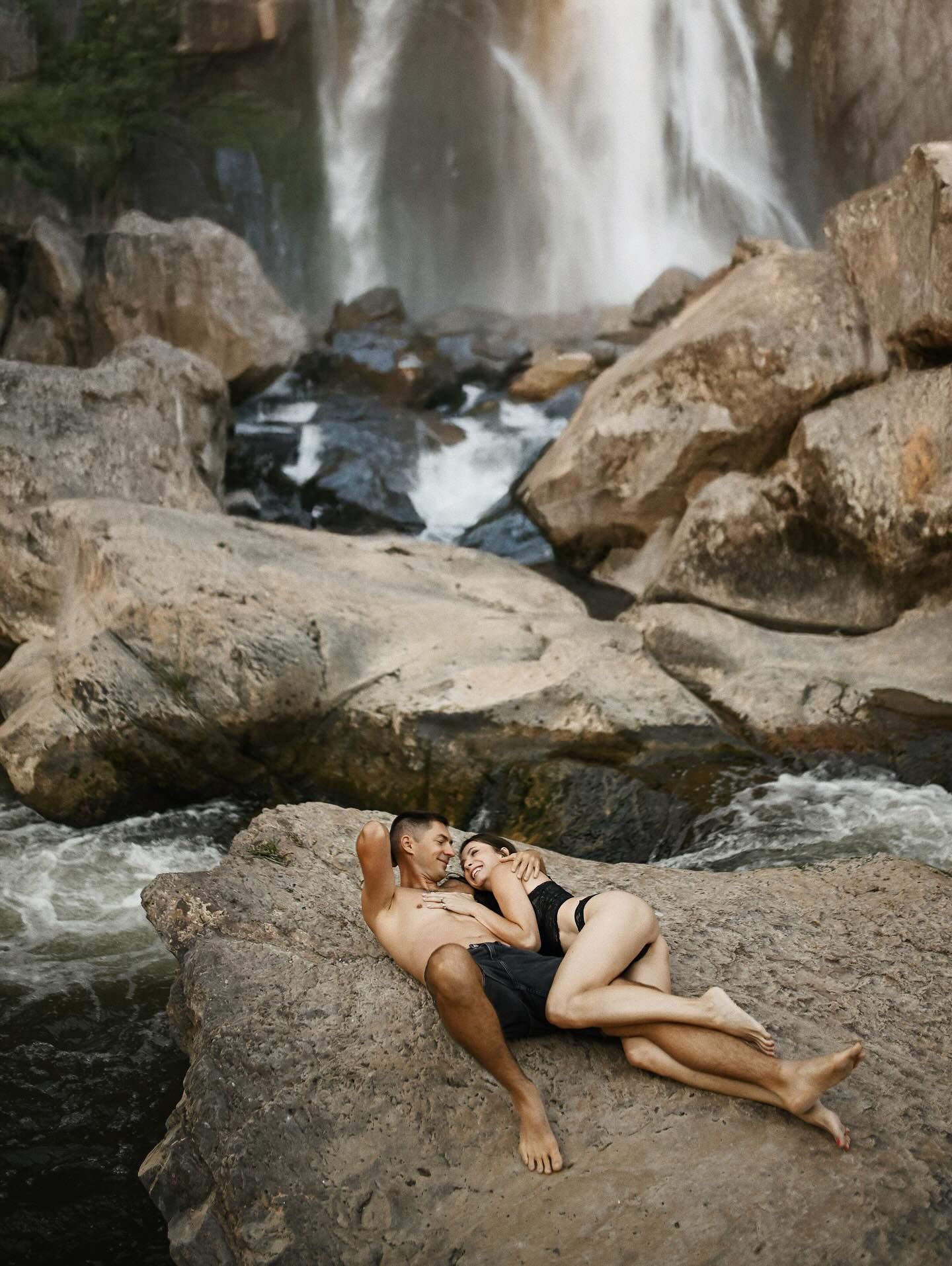 A couple in swimwear relaxing and cuddling closely on stone right next to rushing river stream below a tall natural waterfall near Valencia
