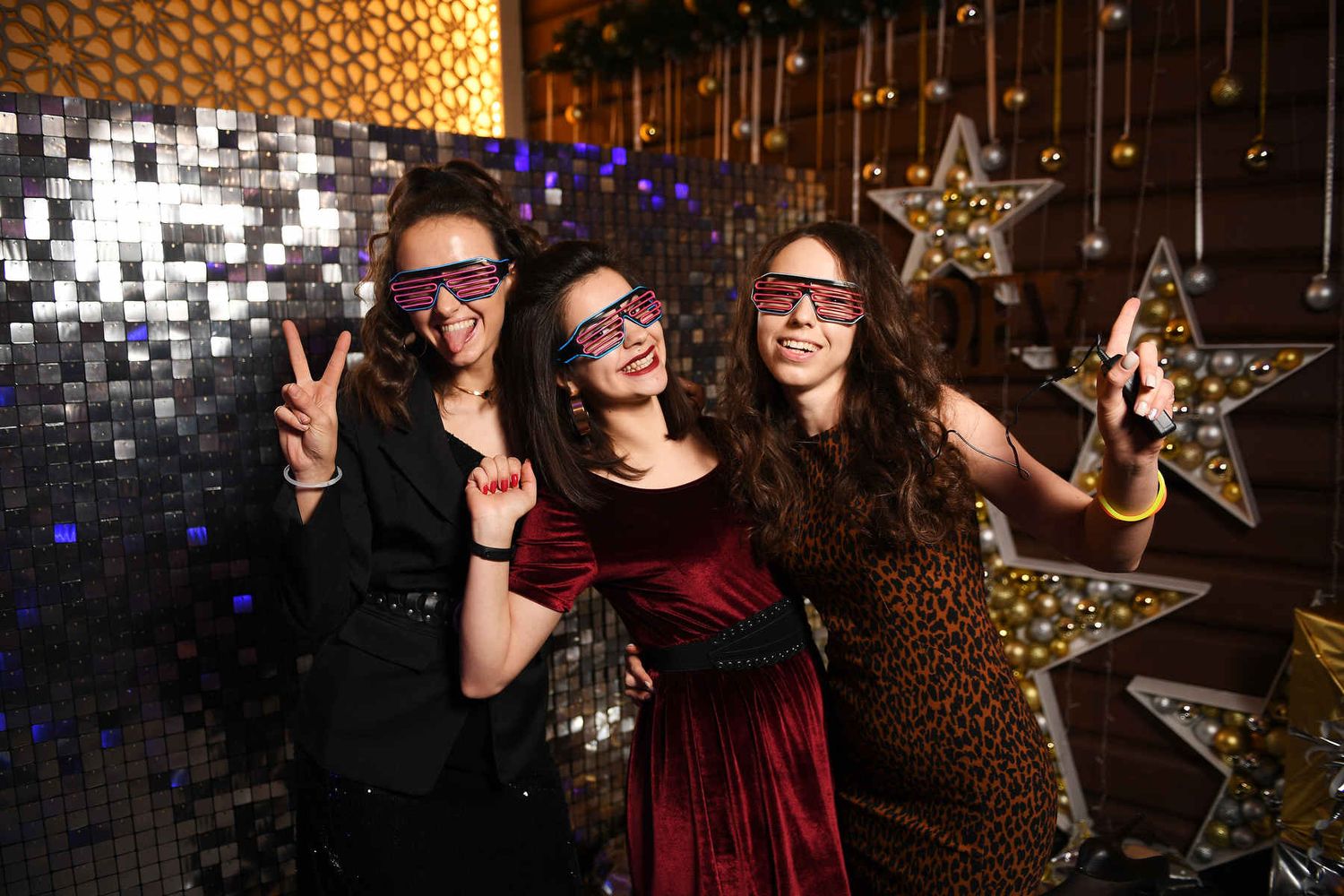 Three happy women wearing futuristic neon LED shutter glasses and posing in front of a sparkling sequin wall.