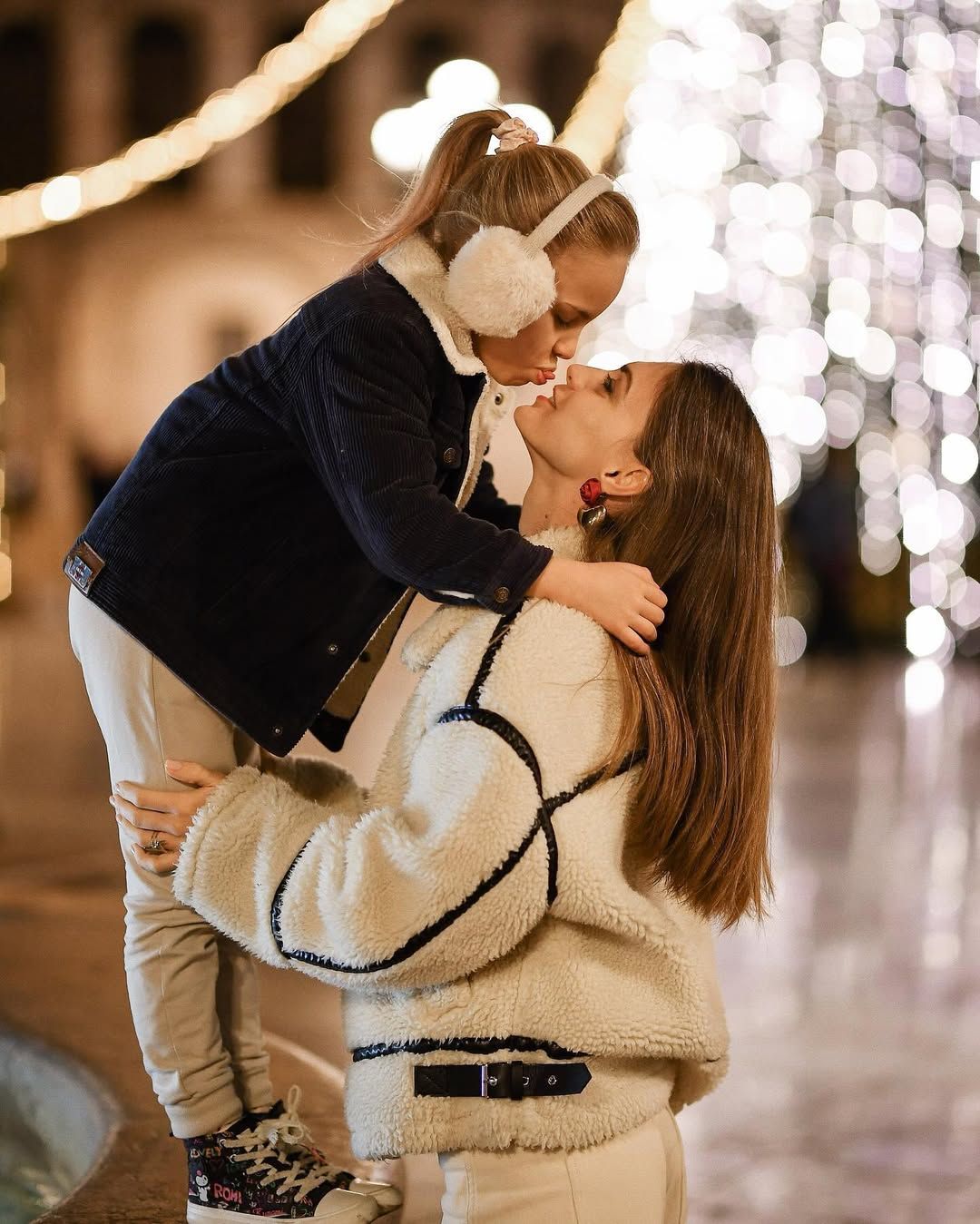 Mother warmly lifting and kissing her little girl with festive holiday bokeh lights in the background in Valencia