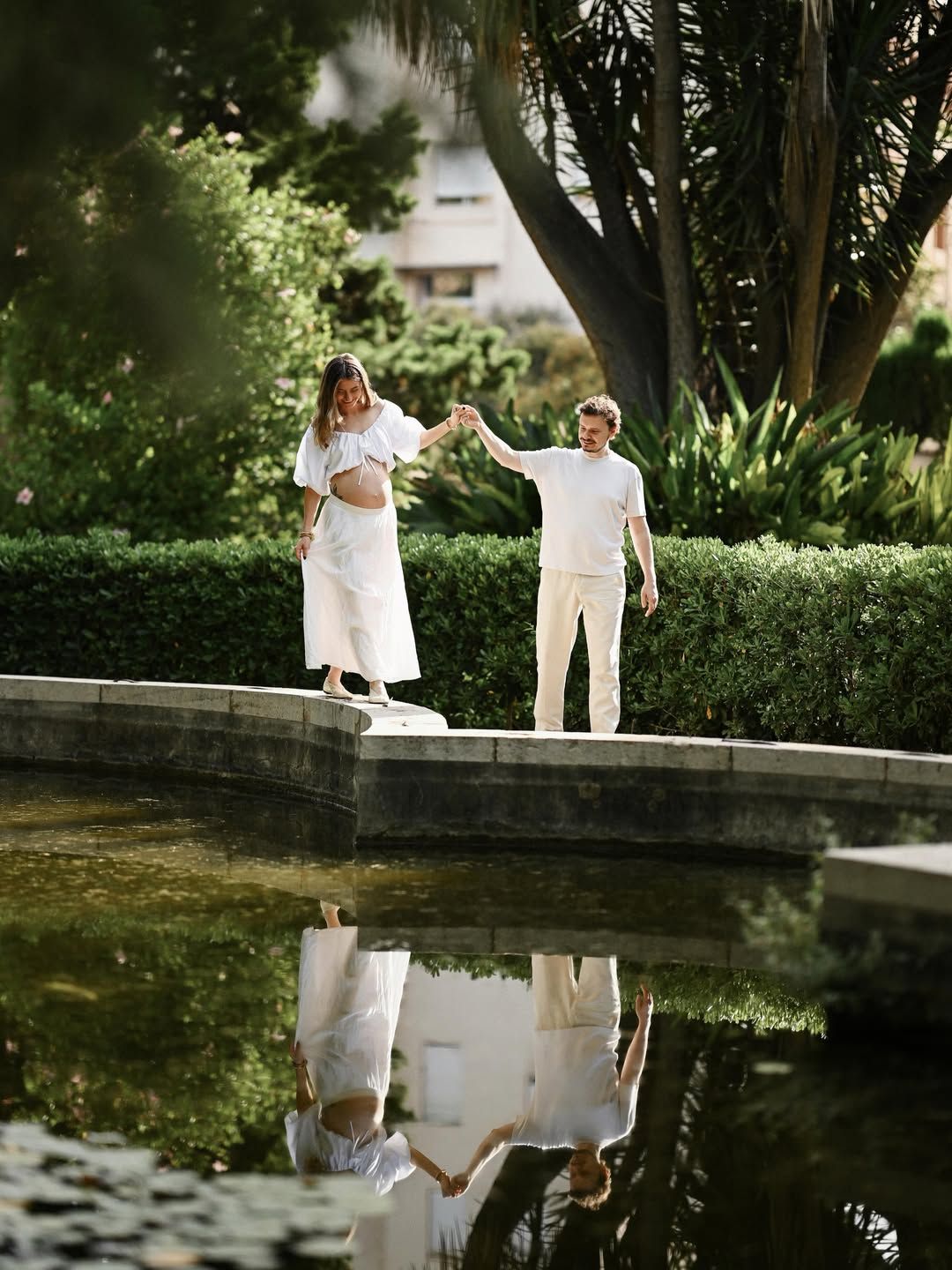 Expecting parents walking on a stone fountain edge with their reflection in the water during a Monforte Gardens maternity session.