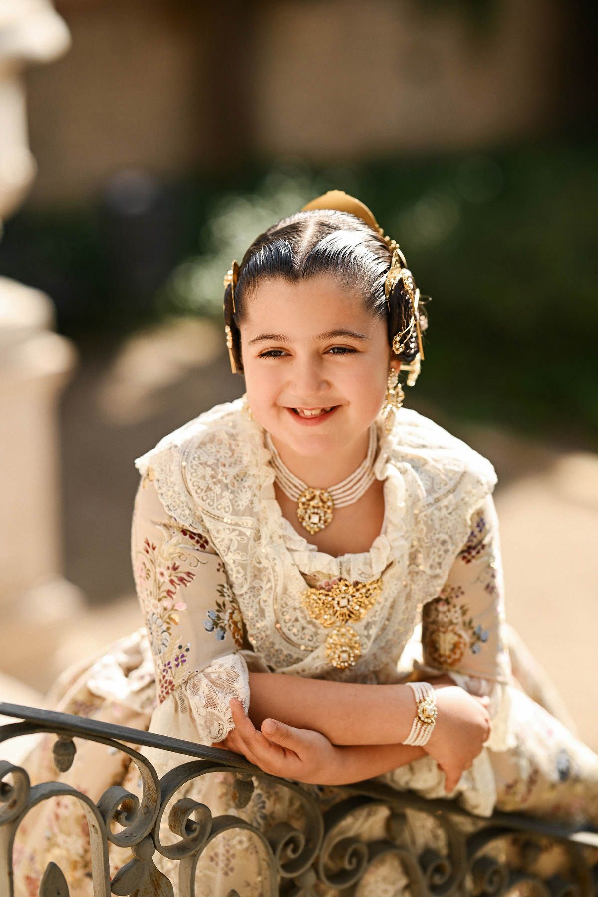 portrait of a smiling young girl in traditional Fallera clothing, posing against an ornate iron railing in the historic Monforte gardens.