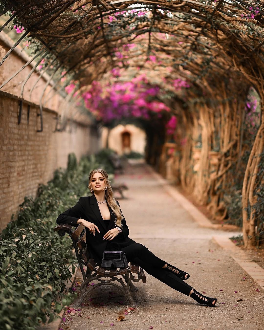A woman in a black outfit sitting on a classic bench under an archway of blooming pink flowers in Valencia.