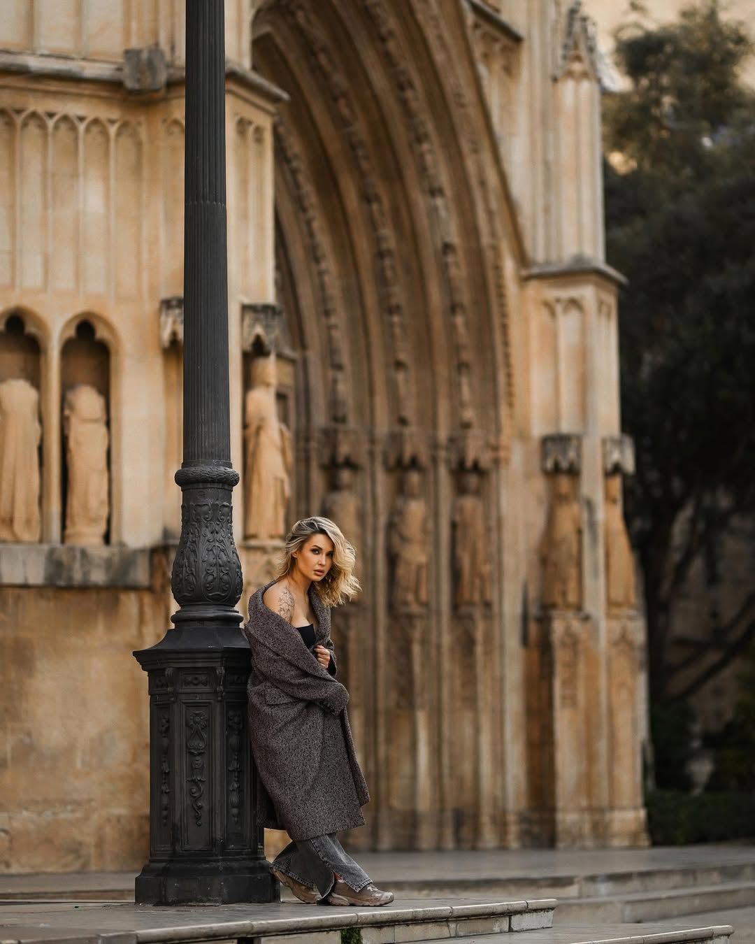 A model in a dark coat leans against a historic lamp post with the majestic gothic arch of the Valencia Cathedral blurred in the background.