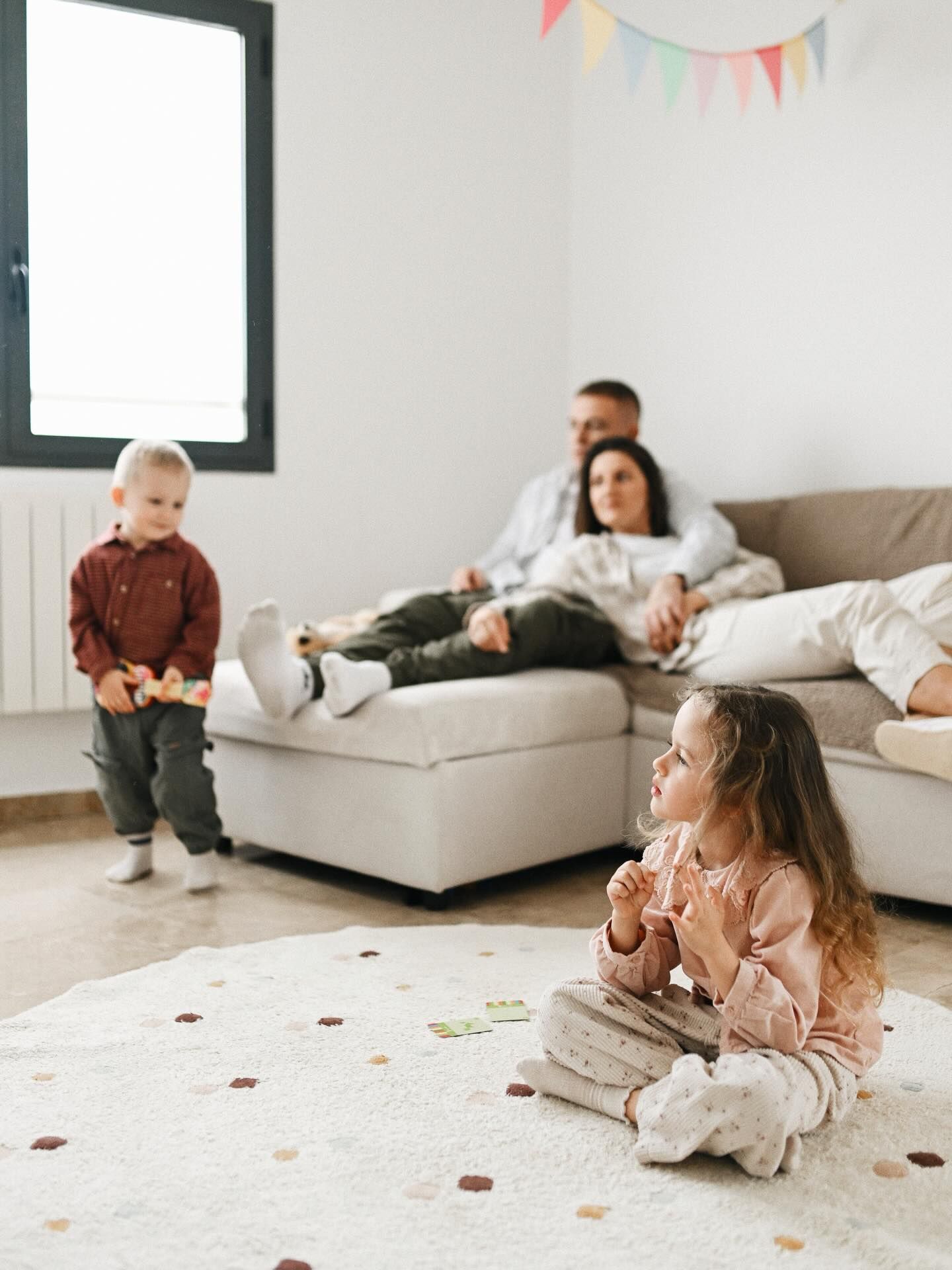 A candid lifestyle home session with parents relaxing on a couch while their two young children play on a rug in the bright living room.
