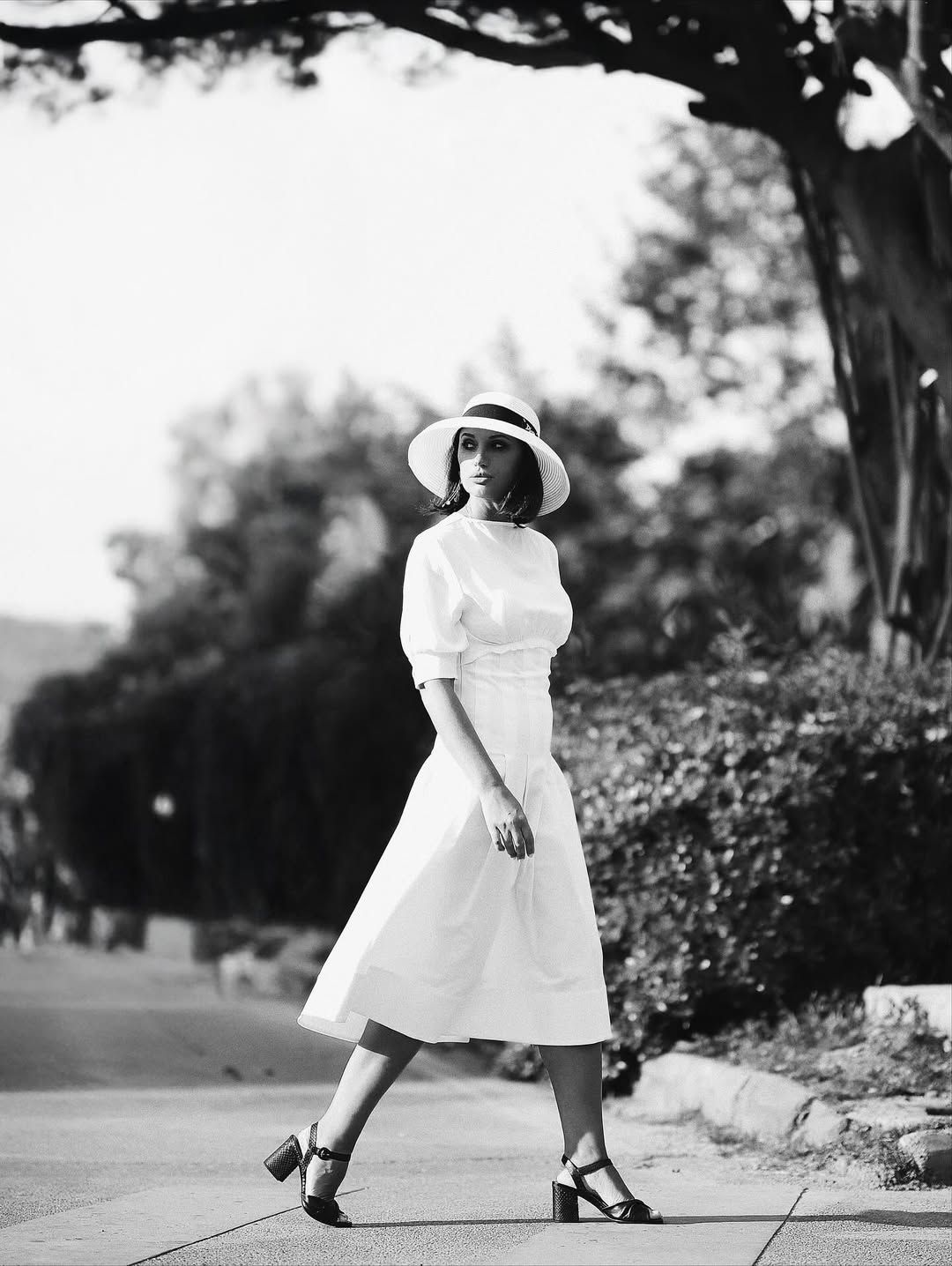 A black and white high-fashion portrait of a woman in a white dress and hat walking down a sunlit pathway in Valencia.