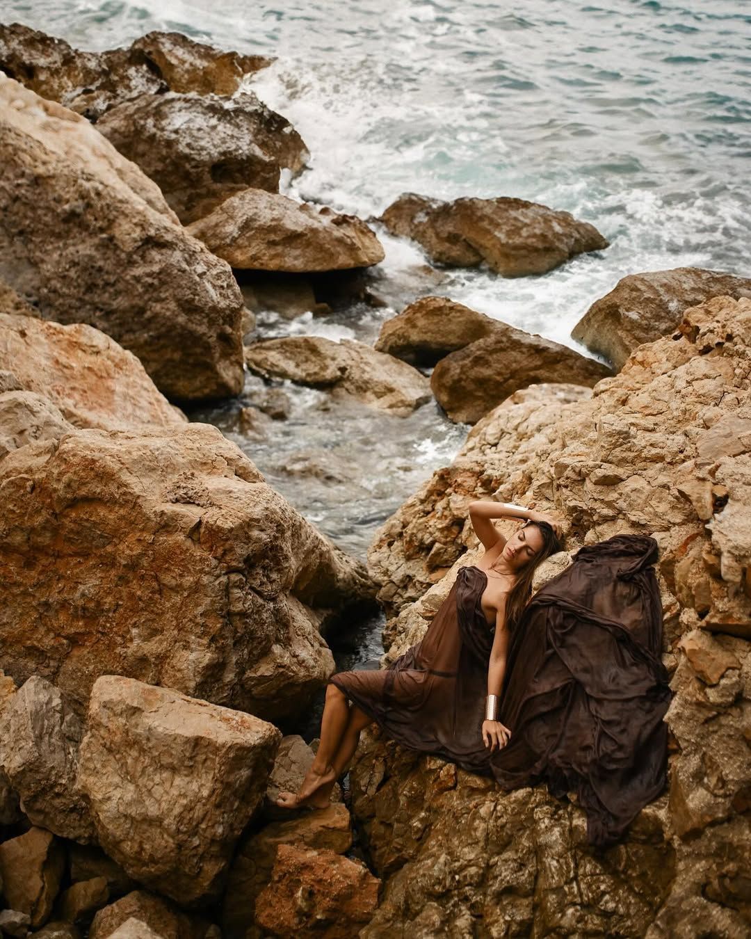 Cinematic portrait of a woman in a sheer dress reclining on rugged rocks at Cala Moraig beach.