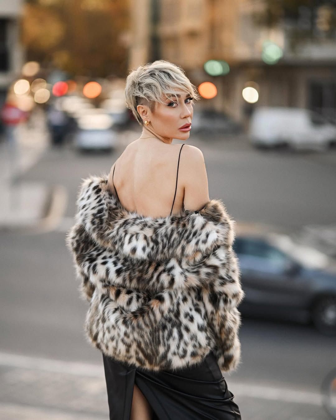 An over-the-shoulder portrait of a luxury styled woman with a short blonde haircut against a blurred city background.