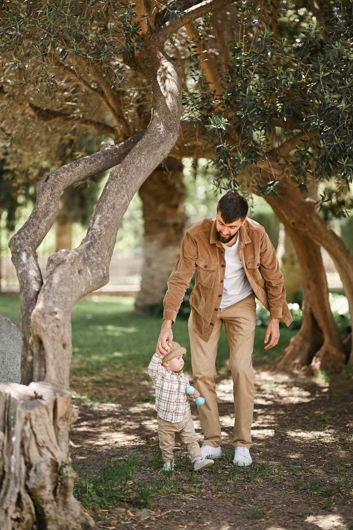 A father gently holding his baby boy's hand to help him walk along a shaded dirt path in Jardines del Real.