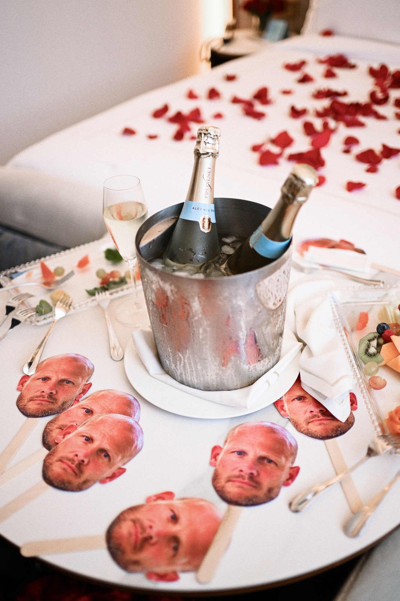 Hen party details in a Valencia hotel room, showing a champagne bucket and funny groom face masks on a table with rose petals.