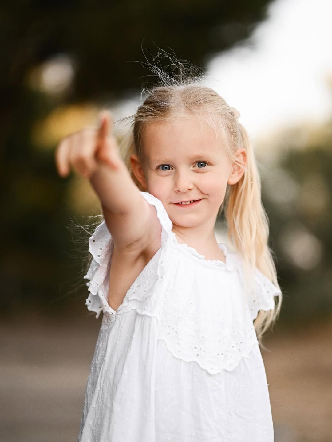 A close-up portrait of a young girl in a white dress pointing toward the camera during a park photoshoot.