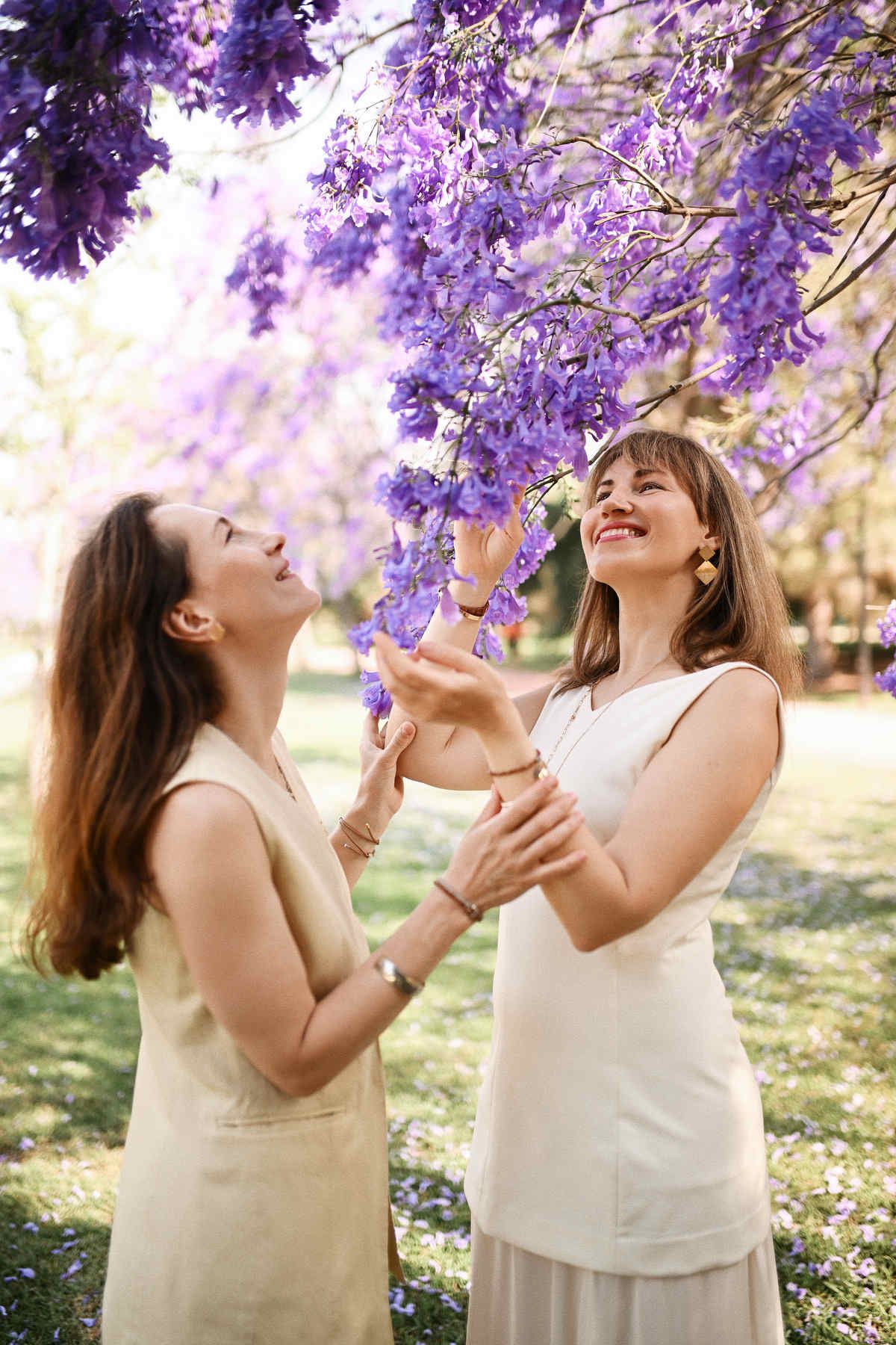 Two elegantly styled women looking up and touching the bright purple flowers of a Jacaranda tree.