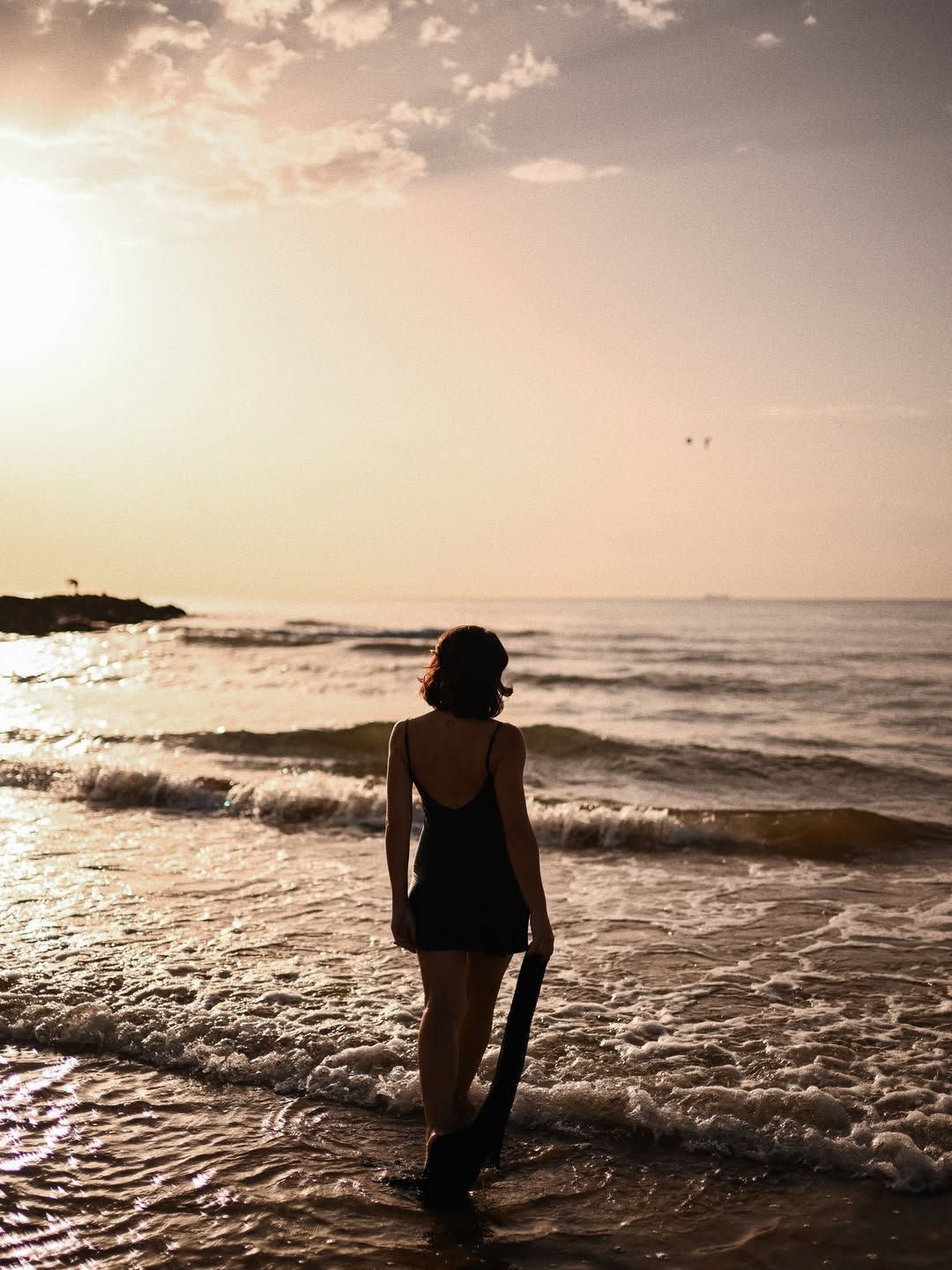 Women's portrait on a Valencia beach, capturing the subject from behind as she wades into the sea waves holding a dark shawl.