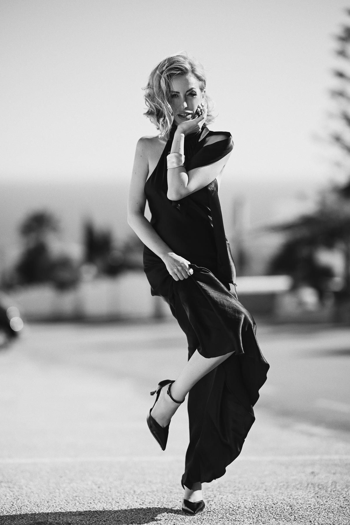 Creative black and white fashion portrait of a woman playfully holding her long black dress while posing on a sunny road