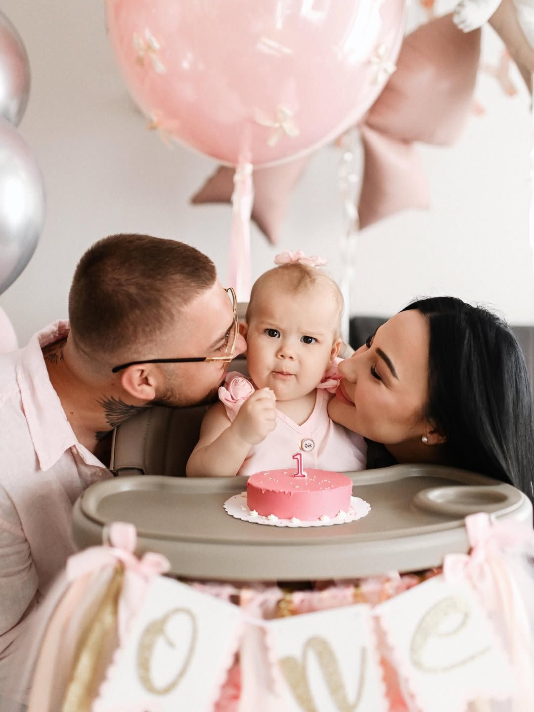 Parents kissing their baby girl sitting in a high chair with a pink birthday cake during an at-home celebration.