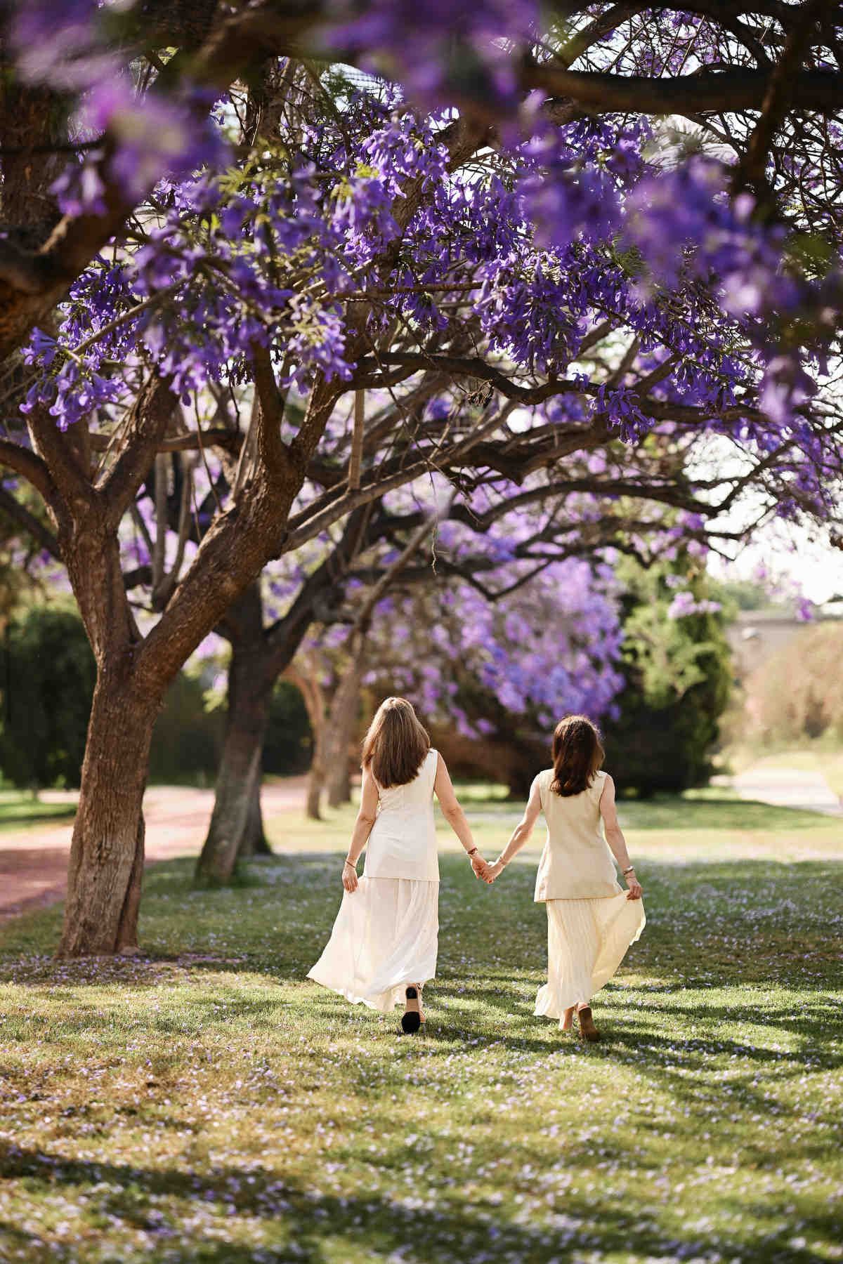 Two adult sisters walking hand in hand along a shaded path under blooming Jacaranda trees.