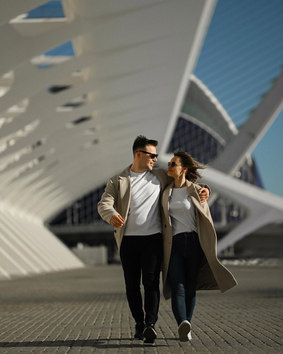 A casual couple wearing beige coats walking and talking under the modern white architectural arches in Valencia.
