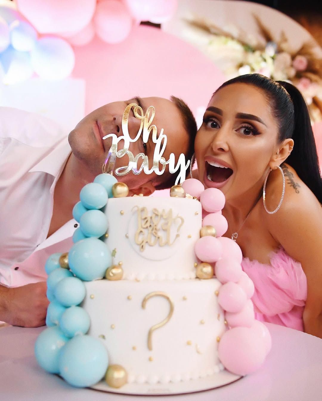 Excited parents-to-be posing behind their custom Oh Baby gender reveal cake during an indoor celebration in Valencia.