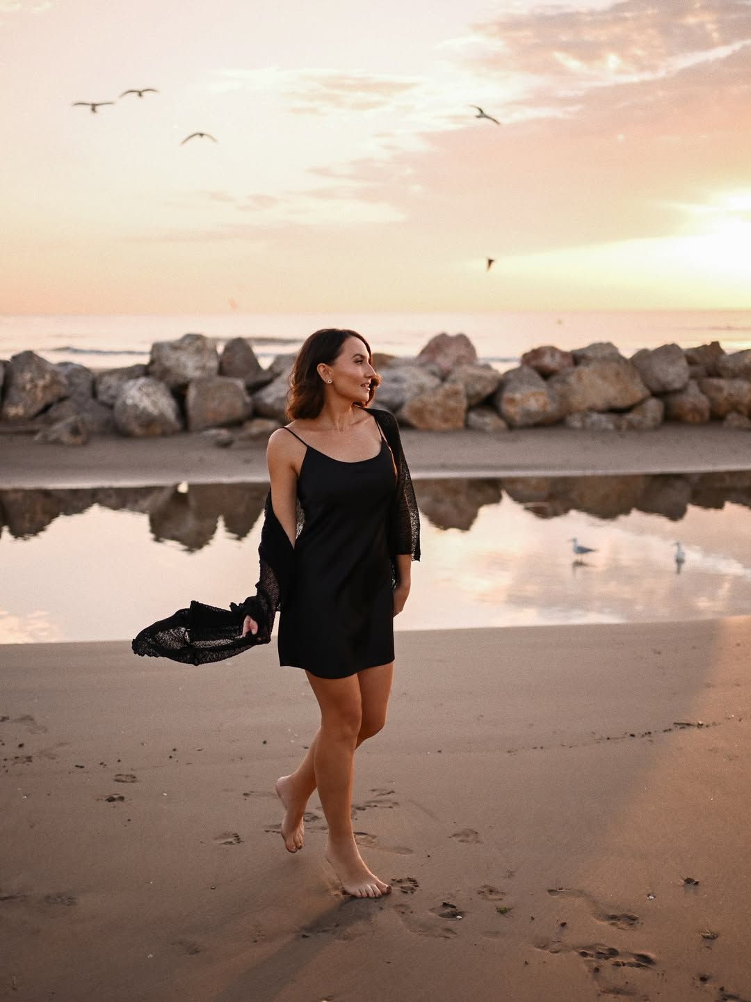 Solo female lifestyle session on a Valencia beach, walking barefoot on wet sand reflecting the pastel sunset sky and seagulls.
