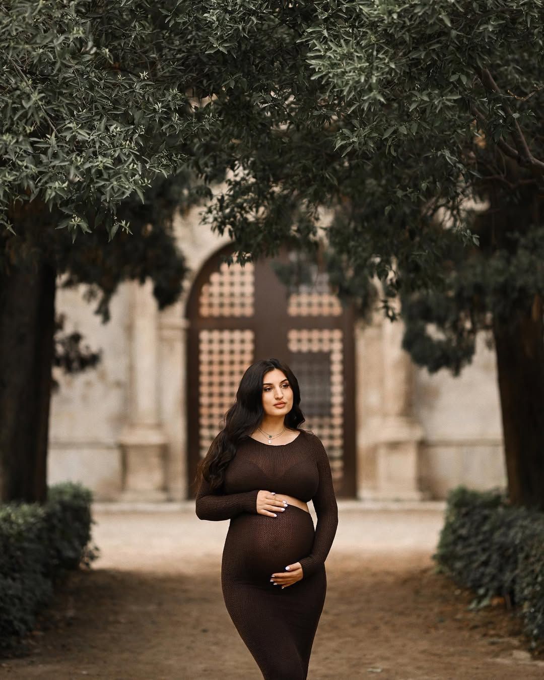 Pregnant woman in a fitted brown dress standing near historic stone architecture at Jardines del Real during a Valencia maternity photoshoot