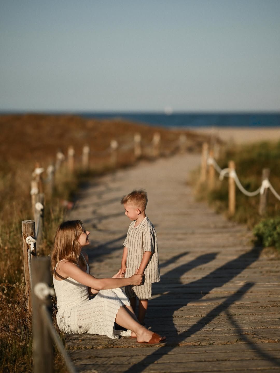 Mother and her young son holding hands on a wooden boardwalk at El Saler beach under warm late afternoon light.