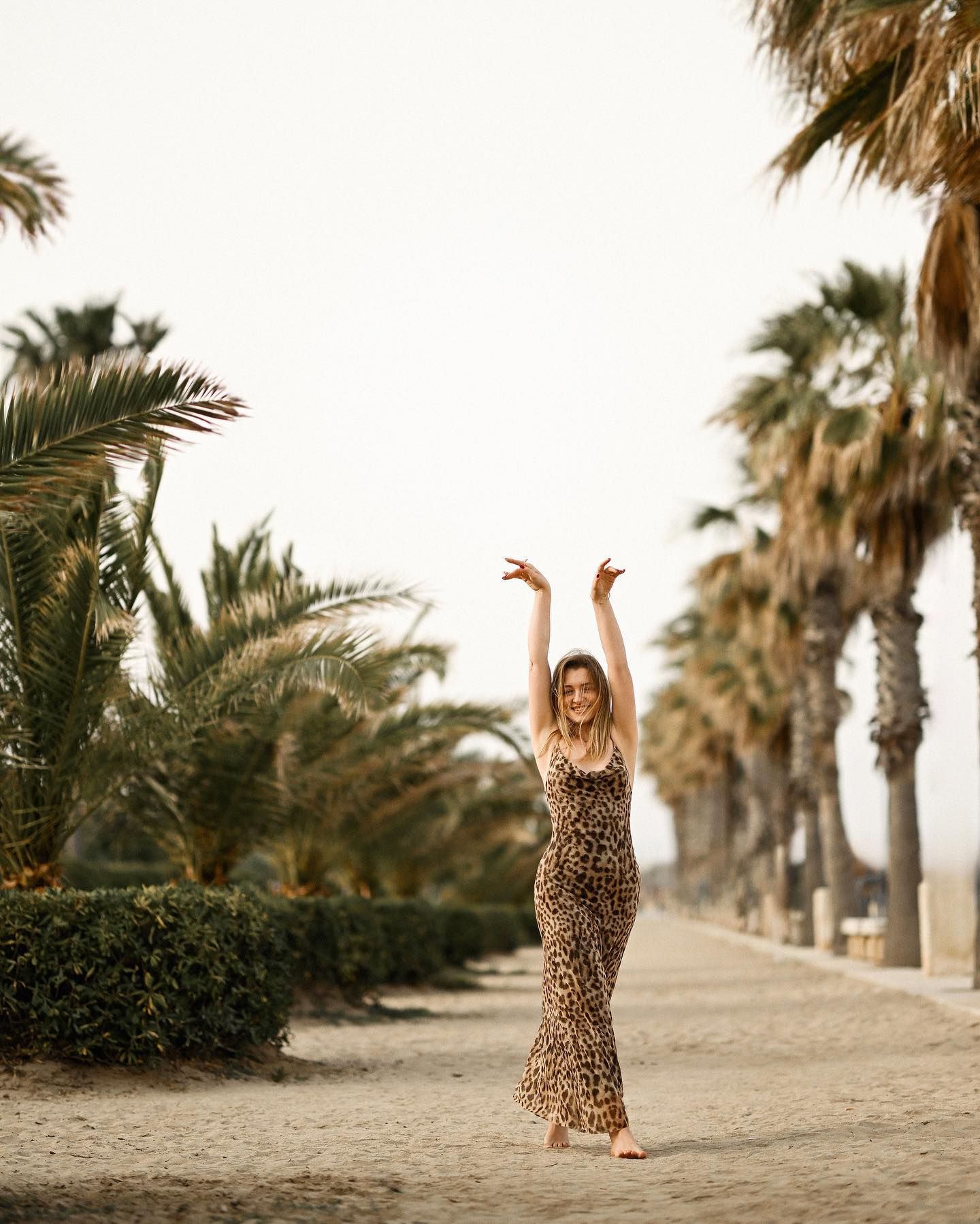 Full-length portrait of a woman in a leopard print dress walking with her arms raised along a palm tree alley in Alicante.