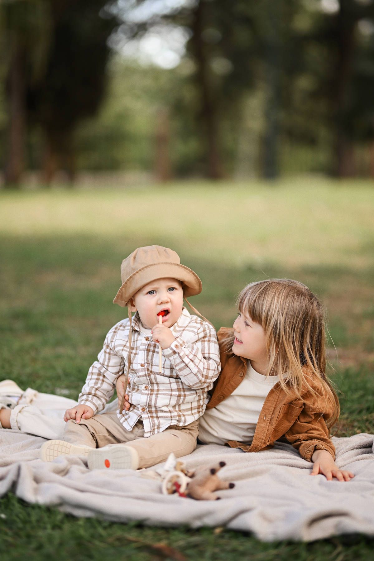 An older sister smiles at her toddler brother holding a red candy while sitting on a blanket on the lawn in Jardines del Real.