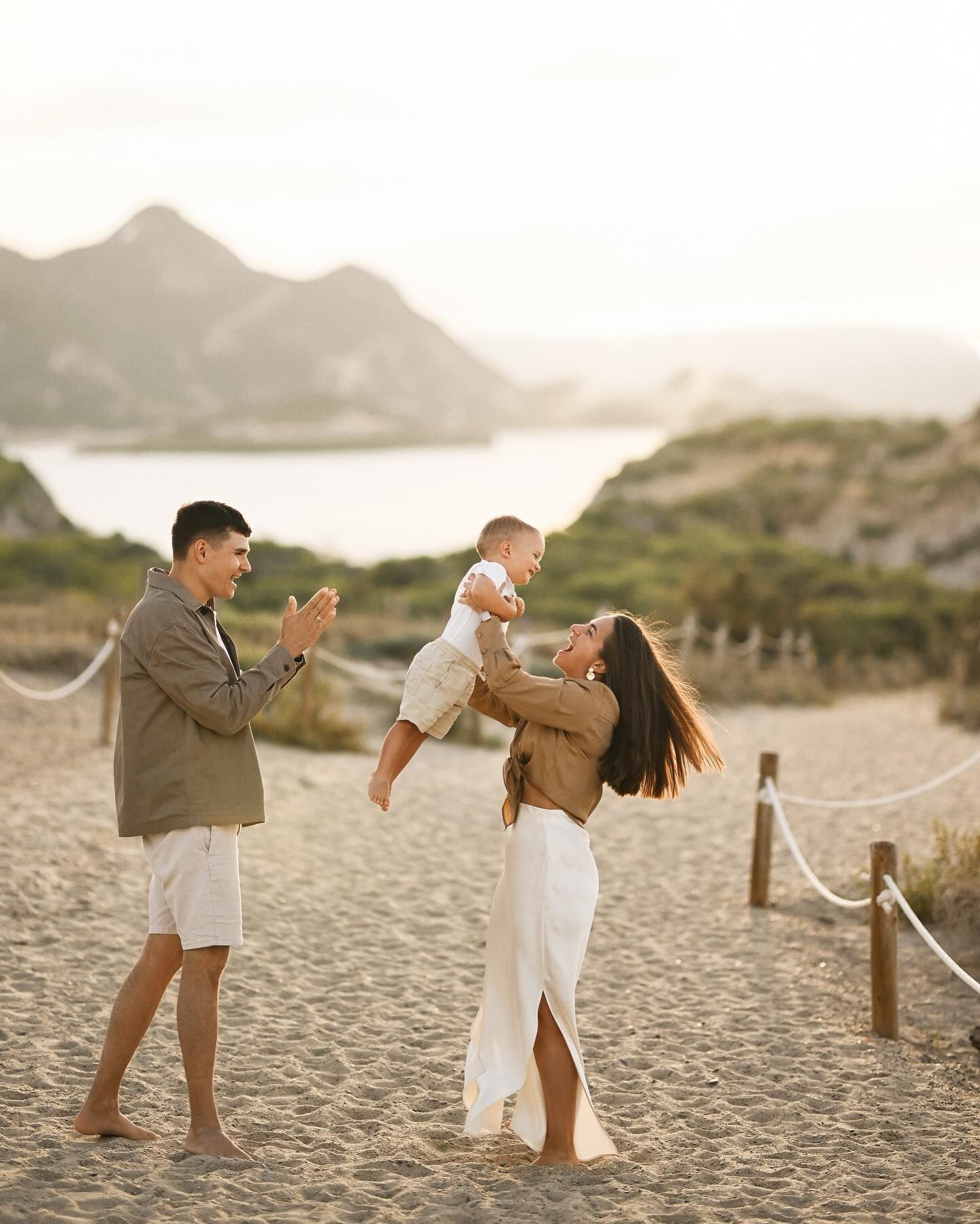 A mother playfully lifting her baby in the air while the father claps, captured at the scenic El Saler beach near Valencia.