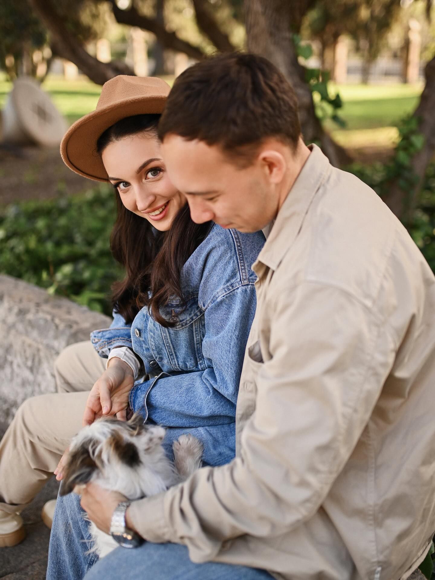 A young woman in a hat looking at the camera while sitting and playing with her small dog and partner on a stone ledge in a park.