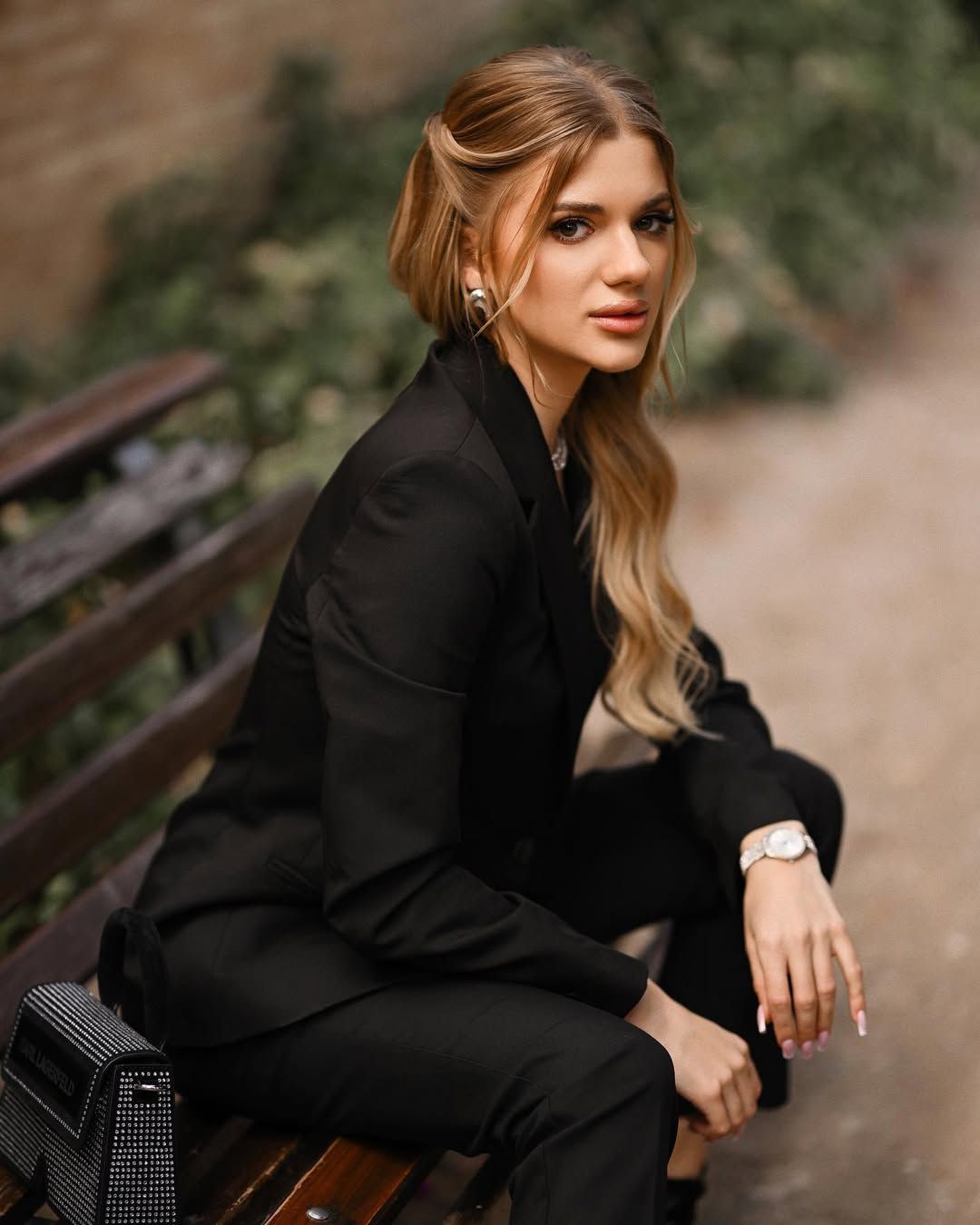 A blonde woman in a black elegant suit sitting on a classic wooden bench in Monforte Gardens.