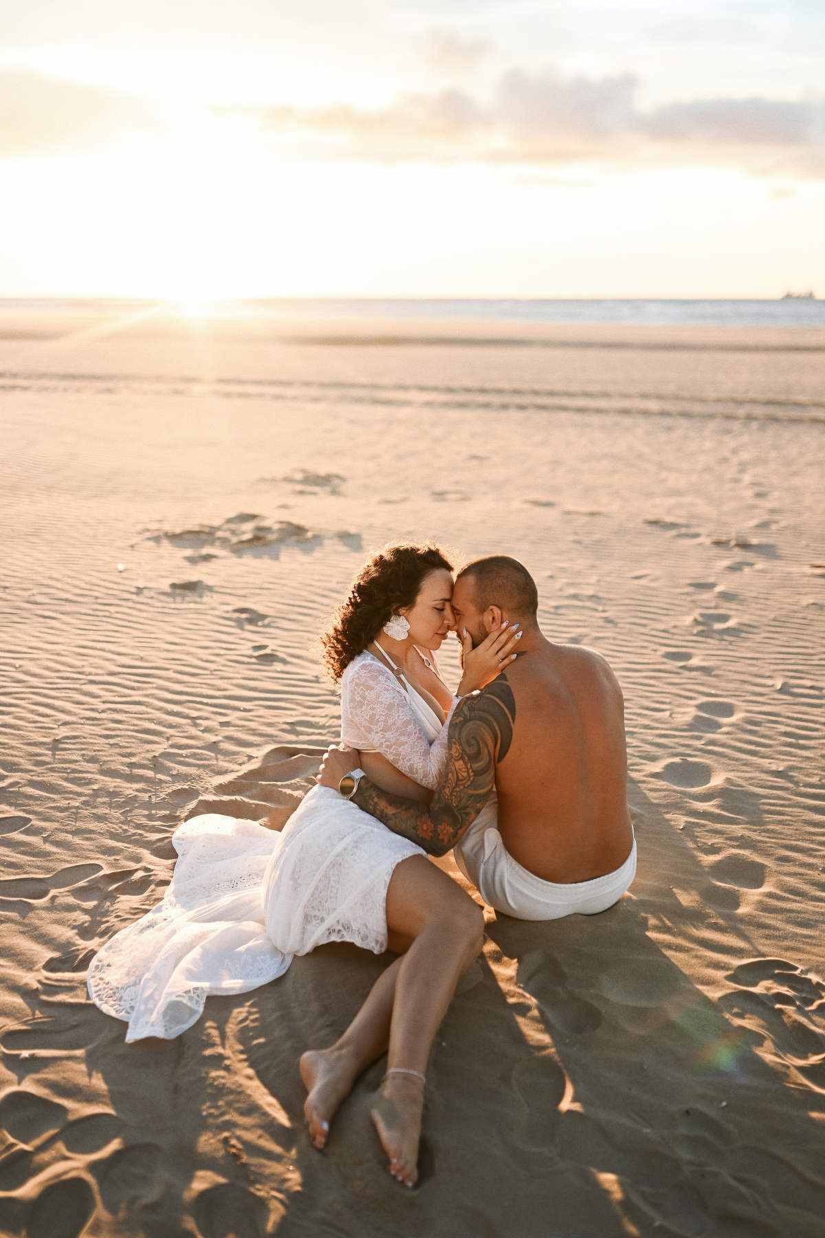 An intimate moment of an expectant couple embracing while sitting on the sand during a golden hour beach session.