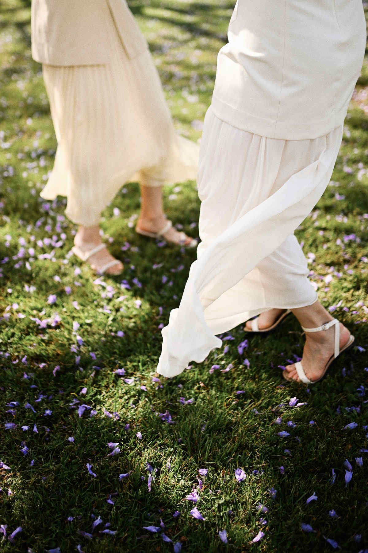 Macro shot of two women in white dresses walking on grass scattered with purple Jacaranda petals.