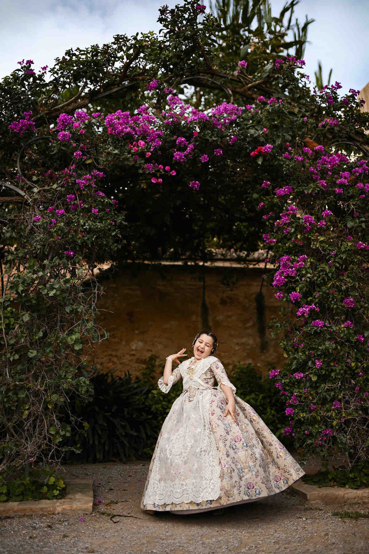 A joyful, candid shot of a young girl in a traditional Fallera dress posing under a blooming bougainvillea arch in Jardines de Monforte.
