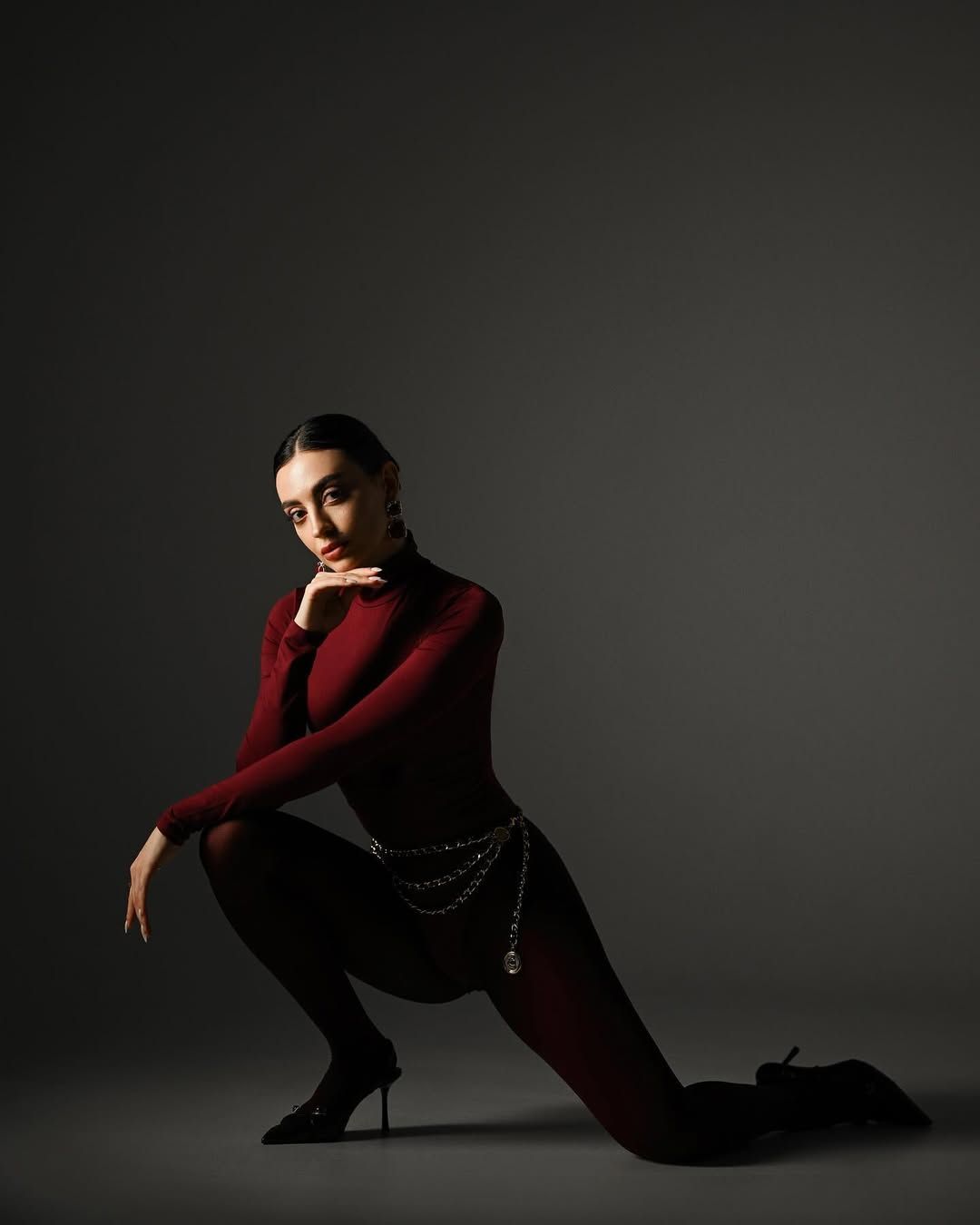 A full-length editorial studio portrait of a woman in a burgundy bodysuit and heels, posing dynamically against a dark grey background.