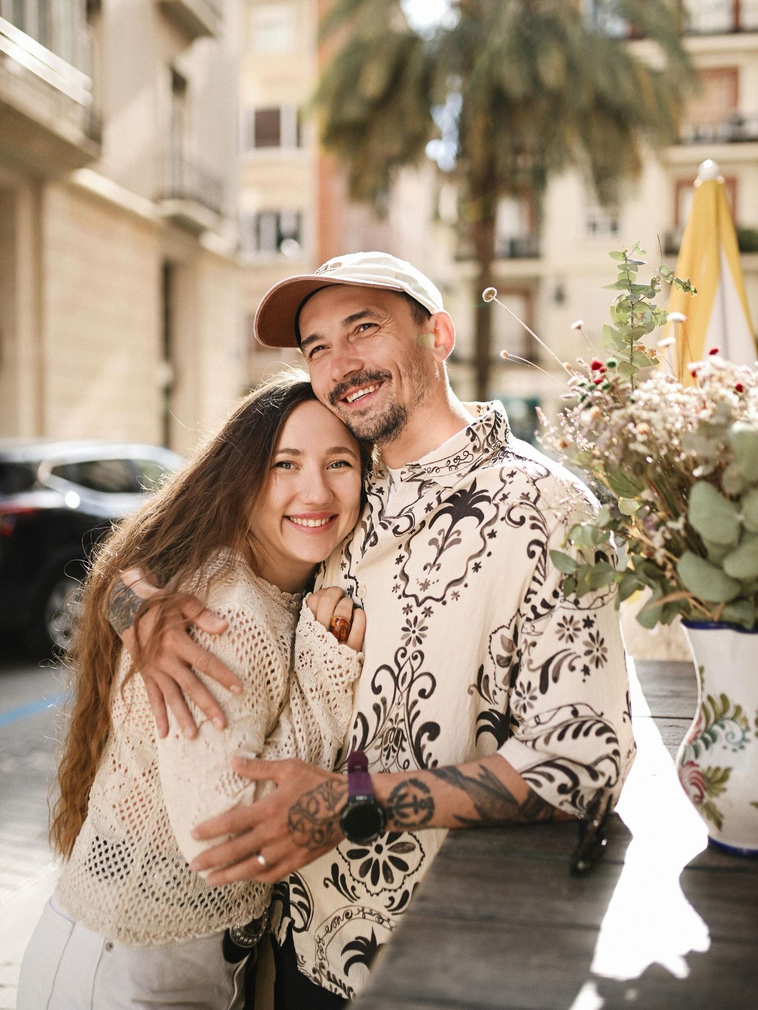A happy couple hugging and smiling warmly at the camera while standing by a decorated table at an outdoor street cafe.