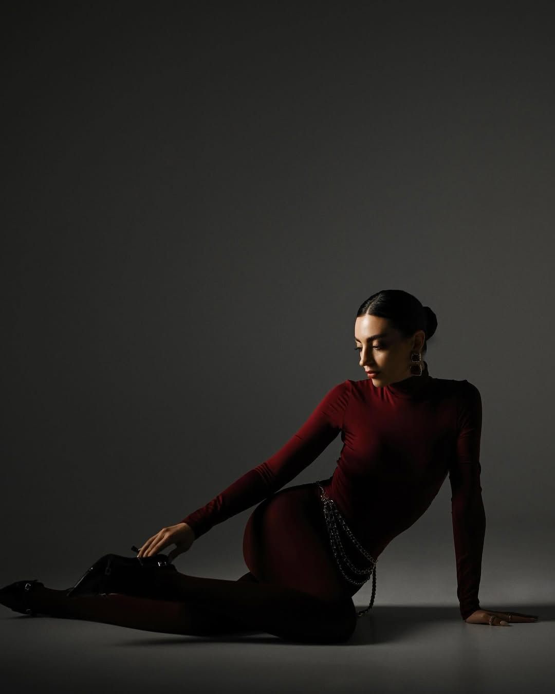 A woman sitting gracefully on the studio floor in a tight dark red outfit, captured in high-contrast editorial lighting.