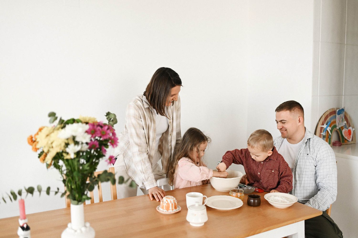 A cheerful family with two children working together to mix ingredients in a bowl at their kitchen table during a home photoshoot.