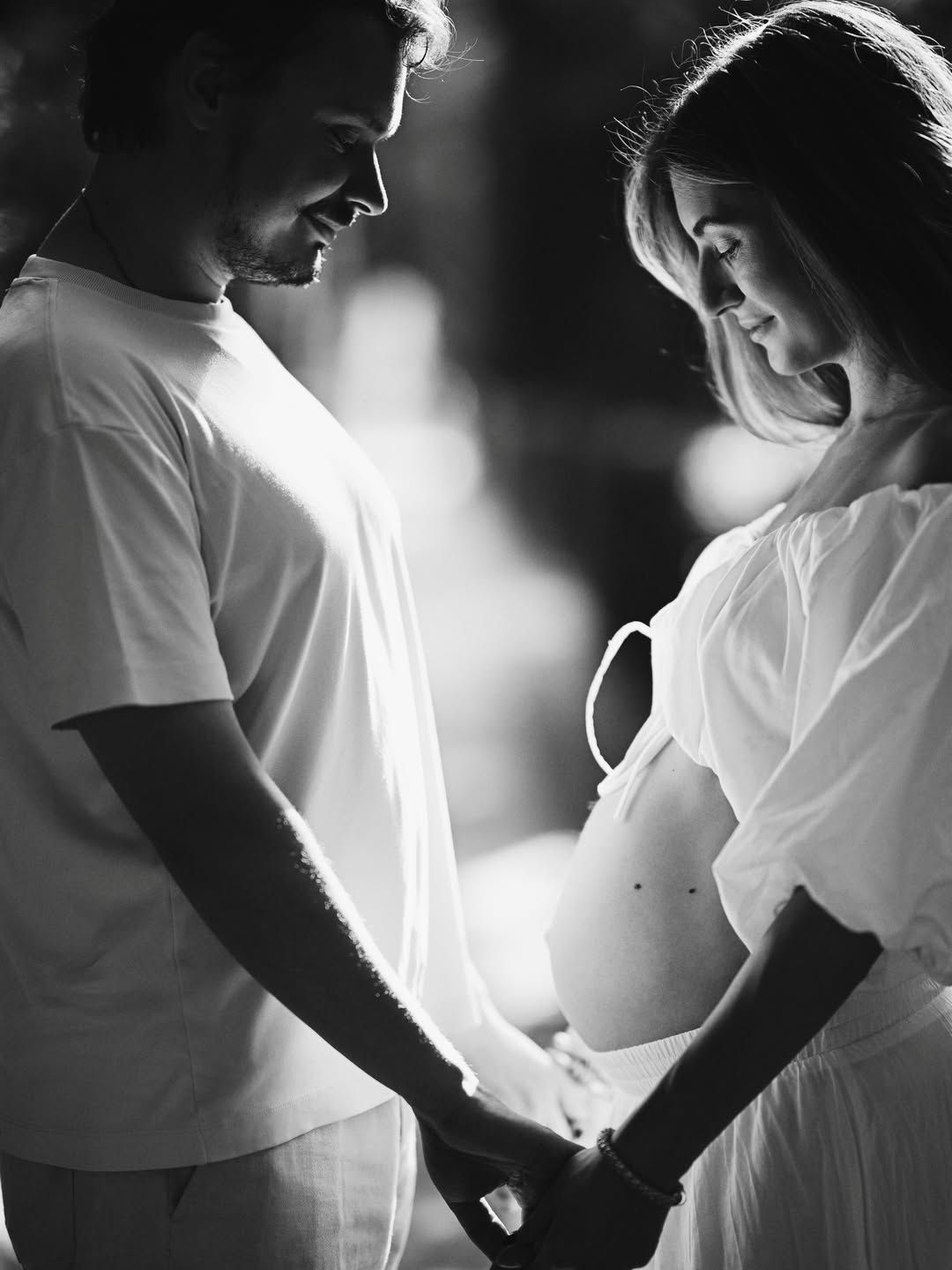 Black and white maternity portrait of a couple holding hands and looking at the pregnancy bump in a quiet Valencia garden