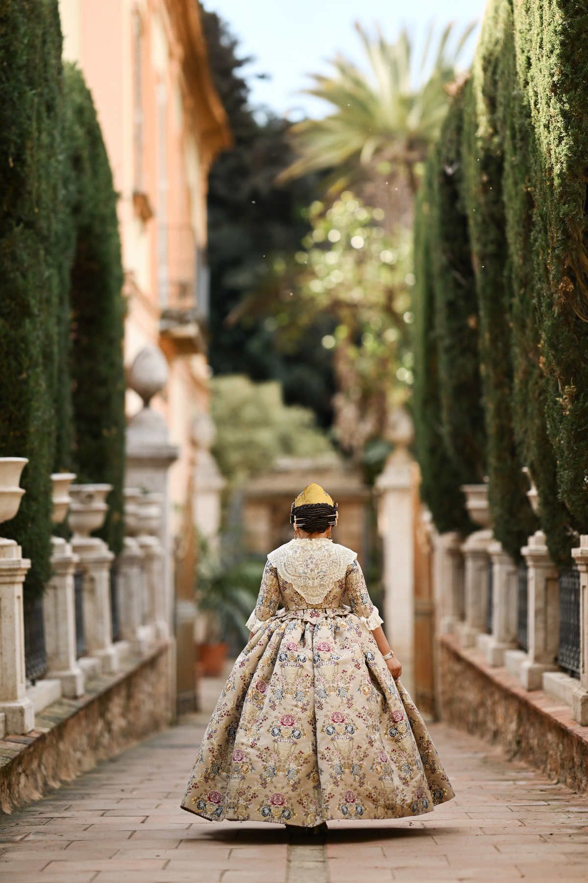 A young girl wearing a traditional traje de fallera dress, showcasing the intricate silk embroidery in the historic Jardines de Monforte.