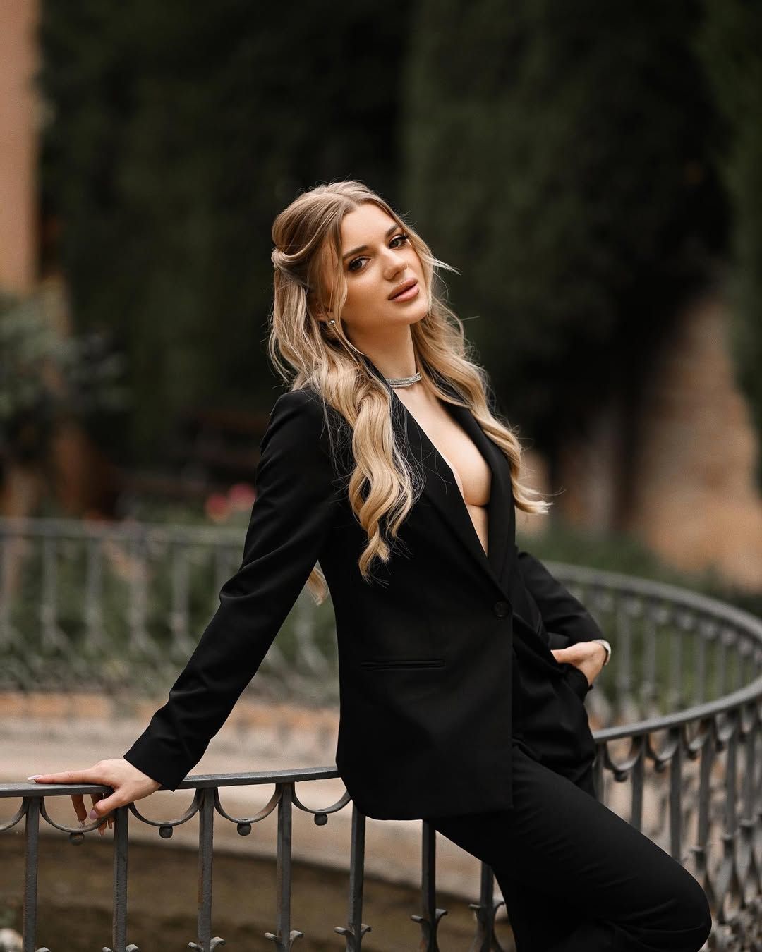 A blonde woman in a structured black pantsuit leaning on a metallic fence in Monforte Gardens, Valencia.