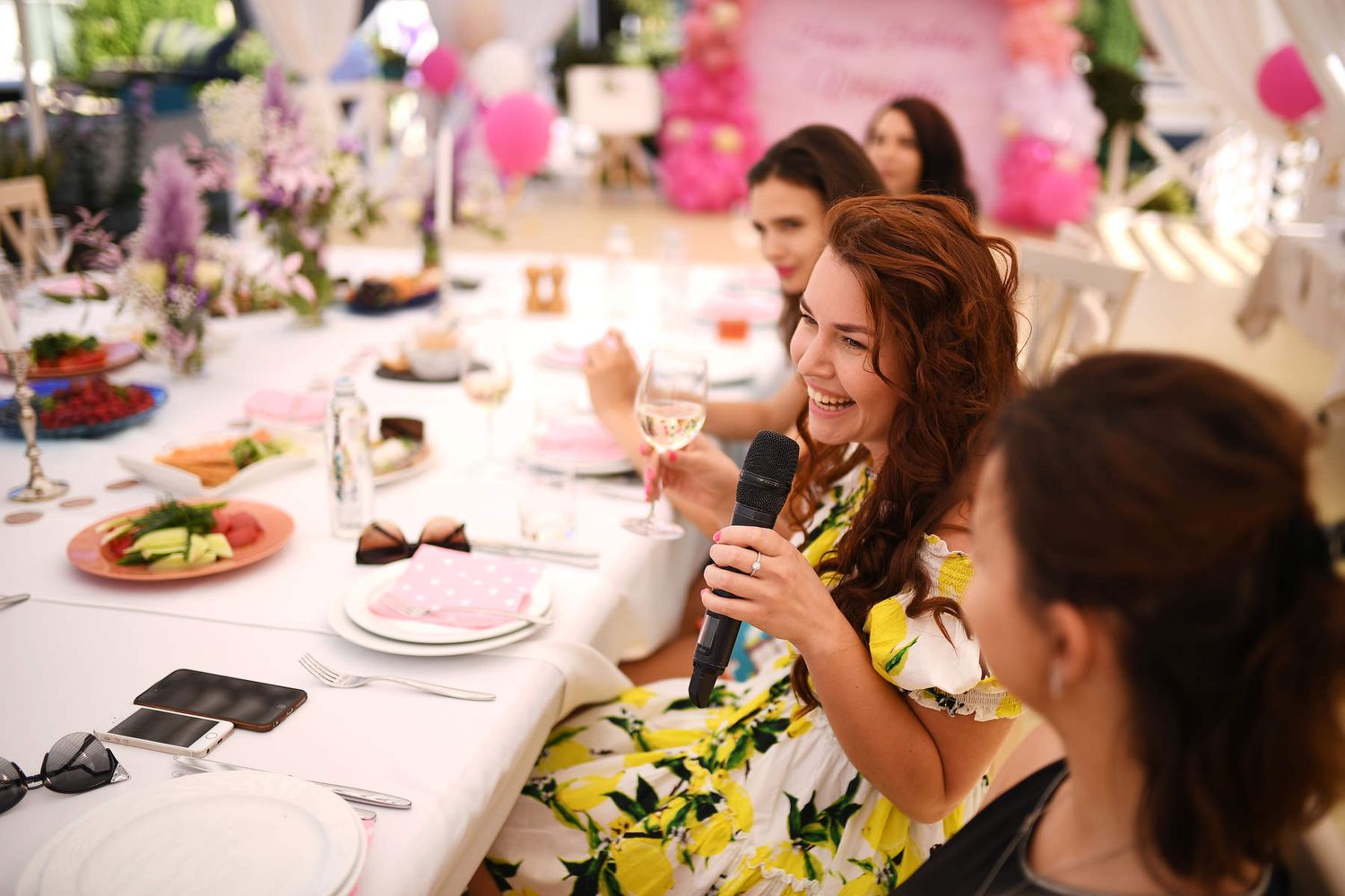 A laughing woman in a lemon-print dress holding a microphone and making a toast during an elegant outdoor banquet.