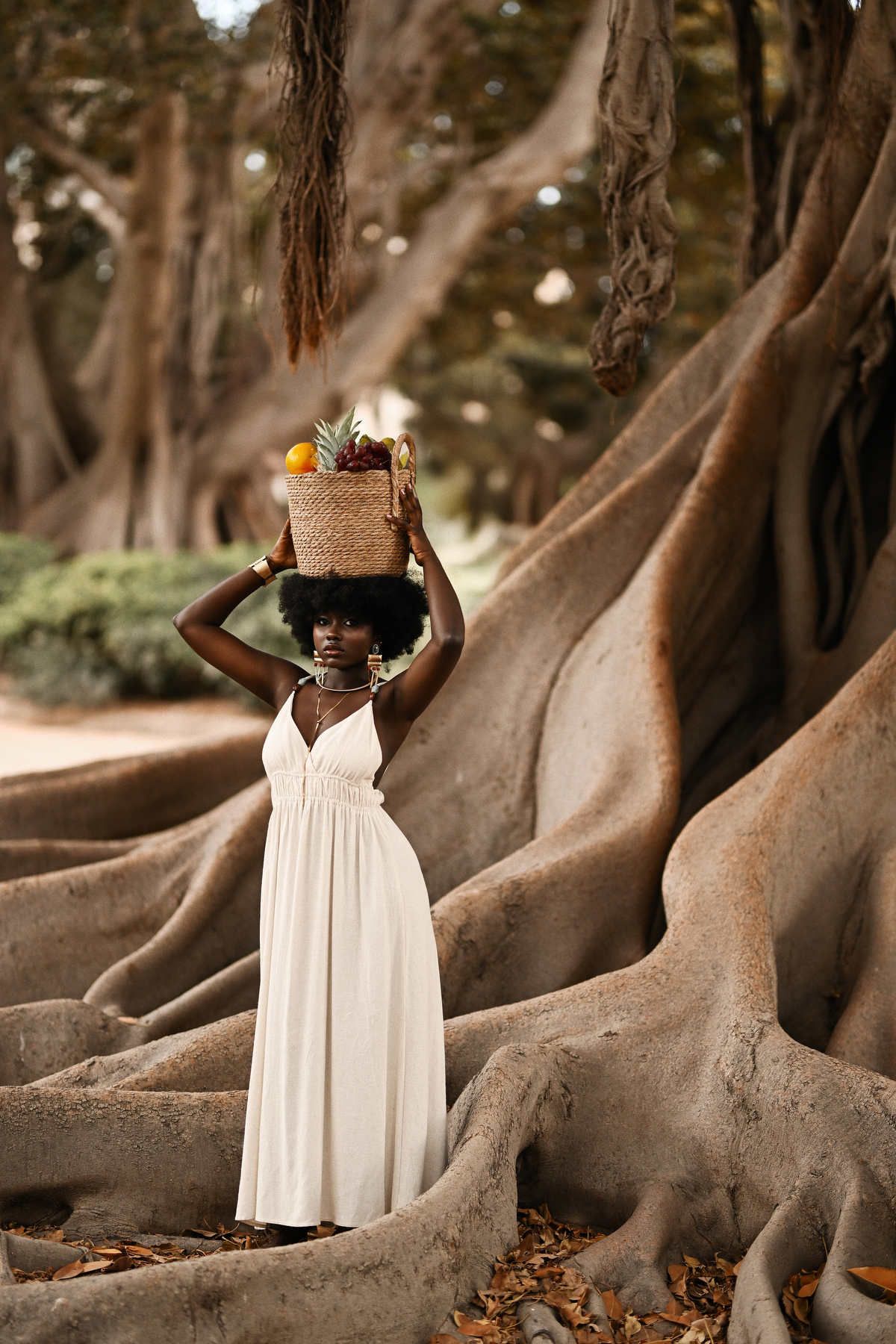 Creative portrait of a model balancing a woven fruit basket on her head while posing by massive roots during a photoshoot in Spain.