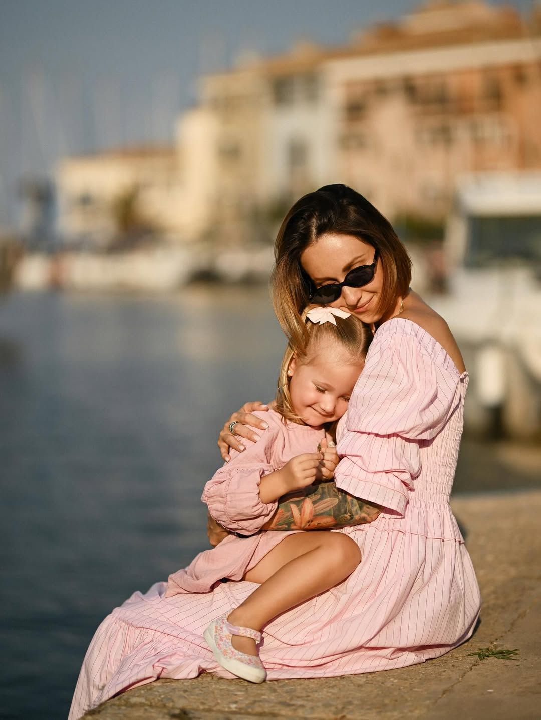 A mother warmly hugging her daughter on the Port Saplaya waterfront promenade with houses and calm water in the background