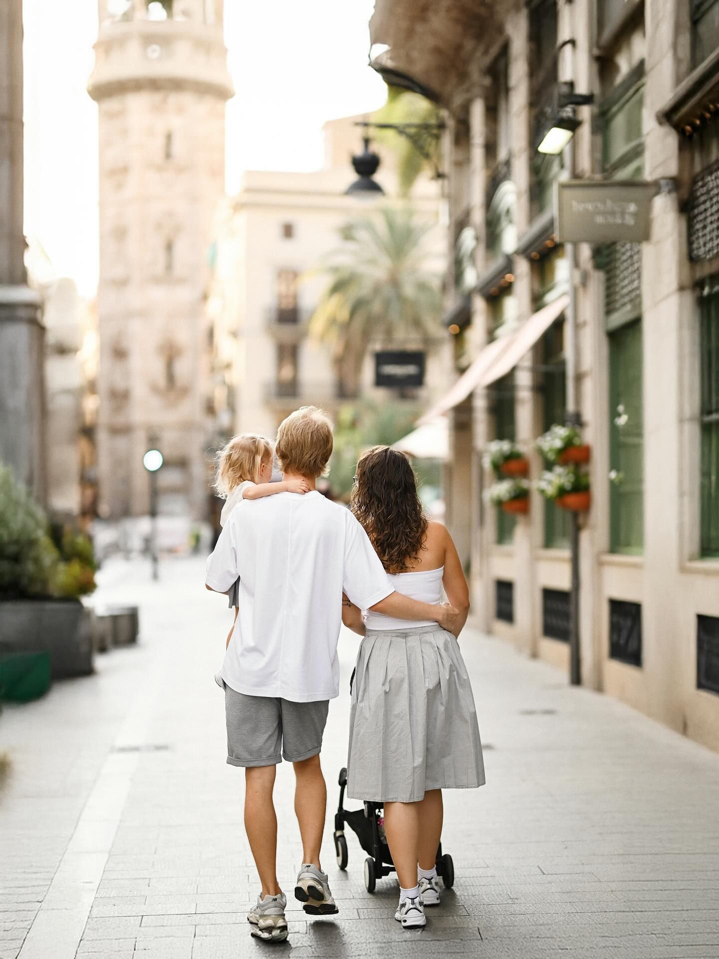 A beautiful family of three walking away from the camera down a scenic Valencian city street while embracing during their photoshoot.