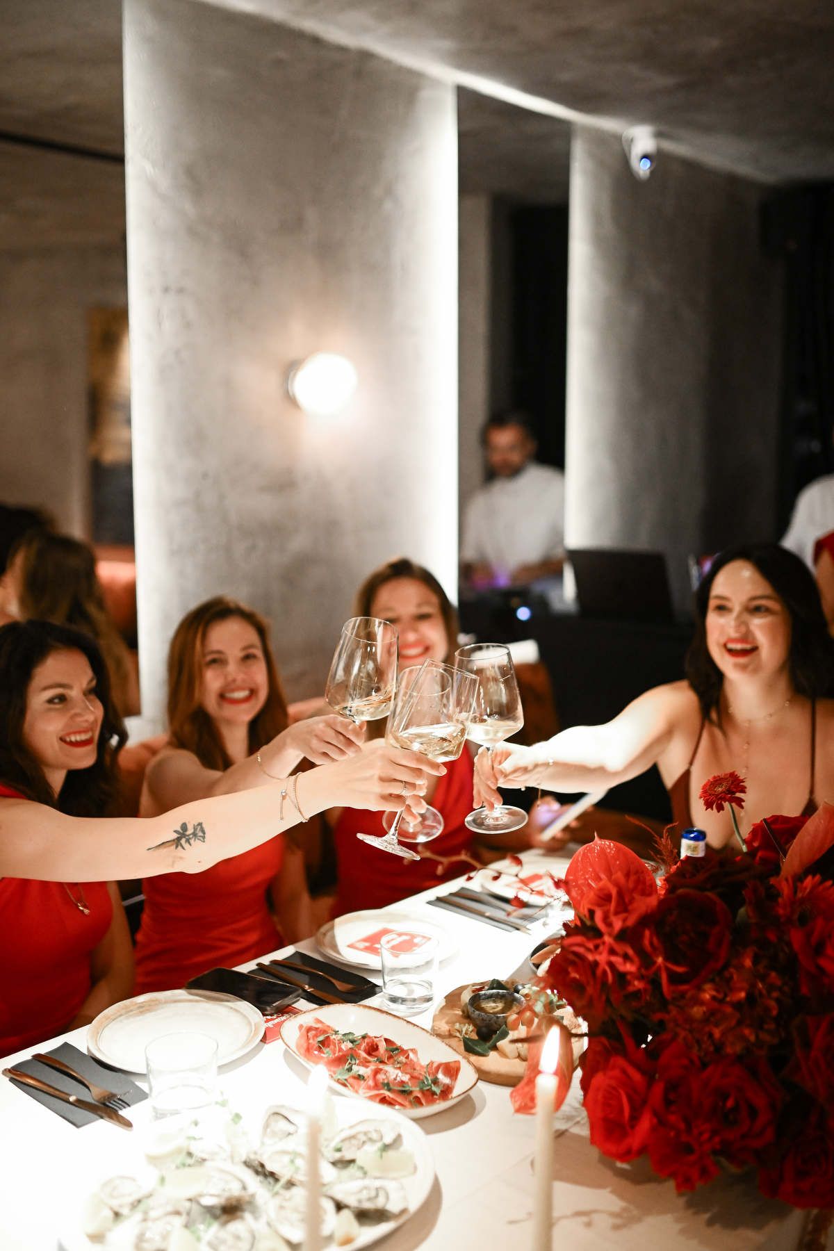 Group of female event guests in matching red dresses clinking wine glasses together during a festive gala dinner.